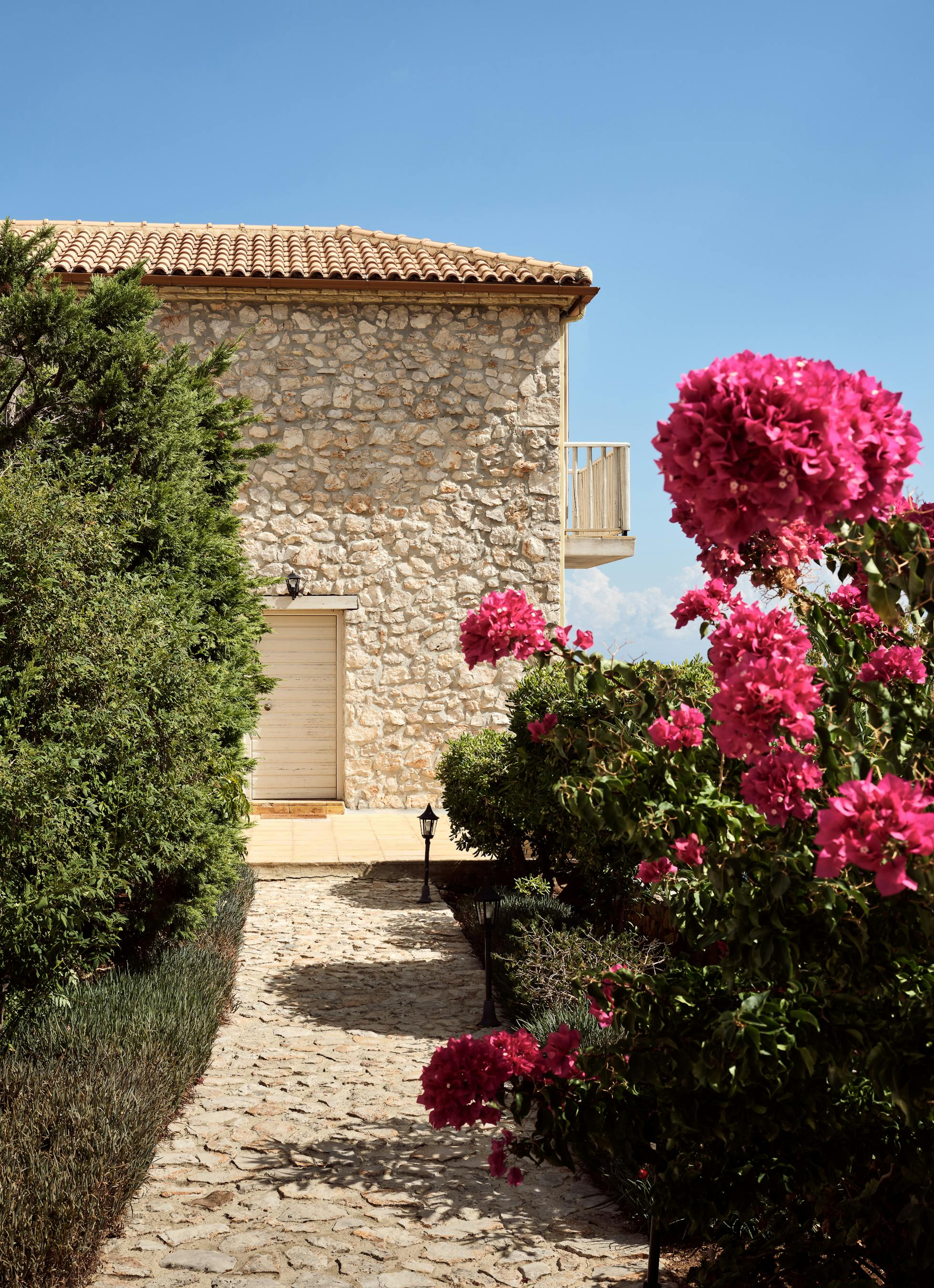 Stone Mediterranean villa entrance with vibrant pink bougainvillea blooms framing a cobblestone pathway leading to a cream-coloured door under terracotta roof tiles.