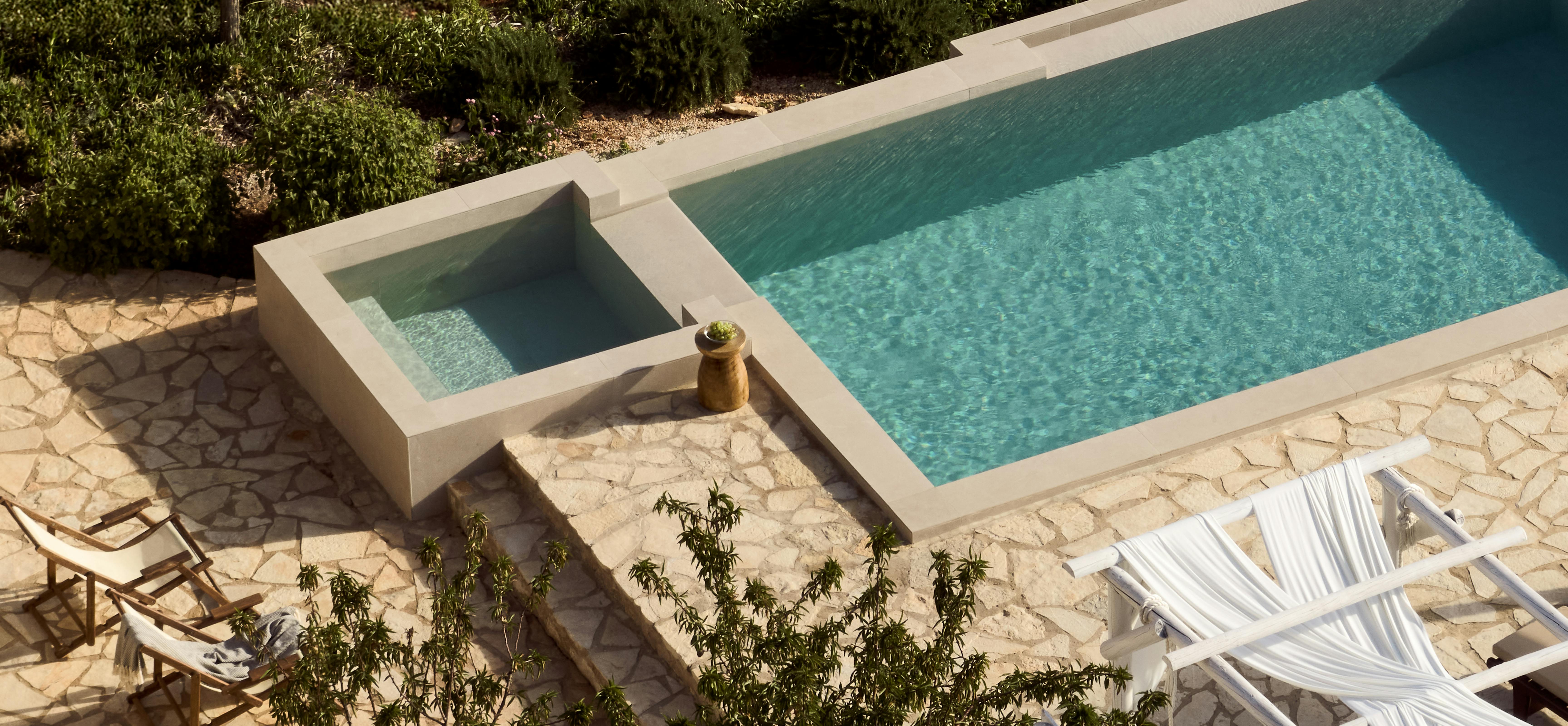 Aerial view of minimalist pool with integrated spa, surrounded by irregular stone paving and white sun loungers beneath trees.