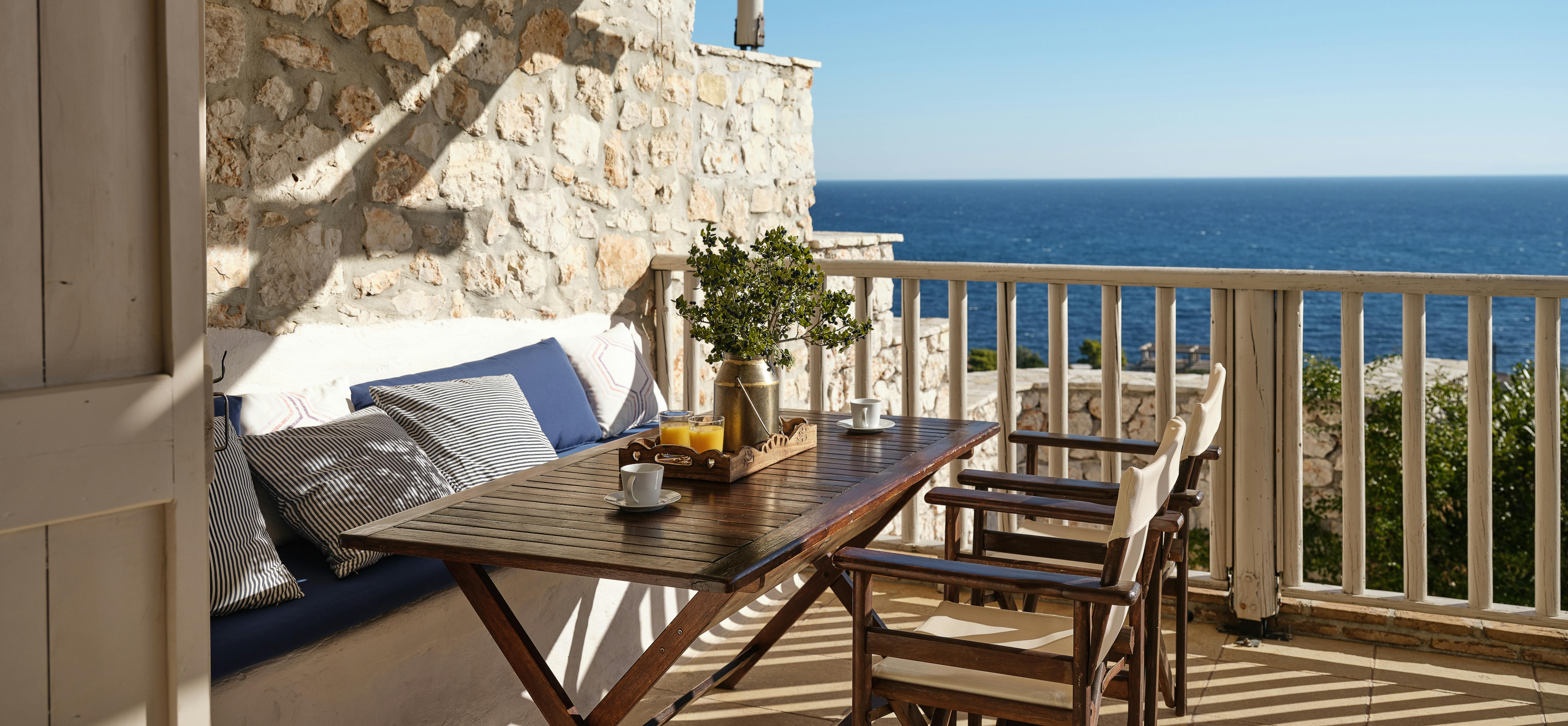 Private balcony with wooden folding table and chairs, built-in bench seating with striped cushions, and white railing overlooking Mediterranean Sea.