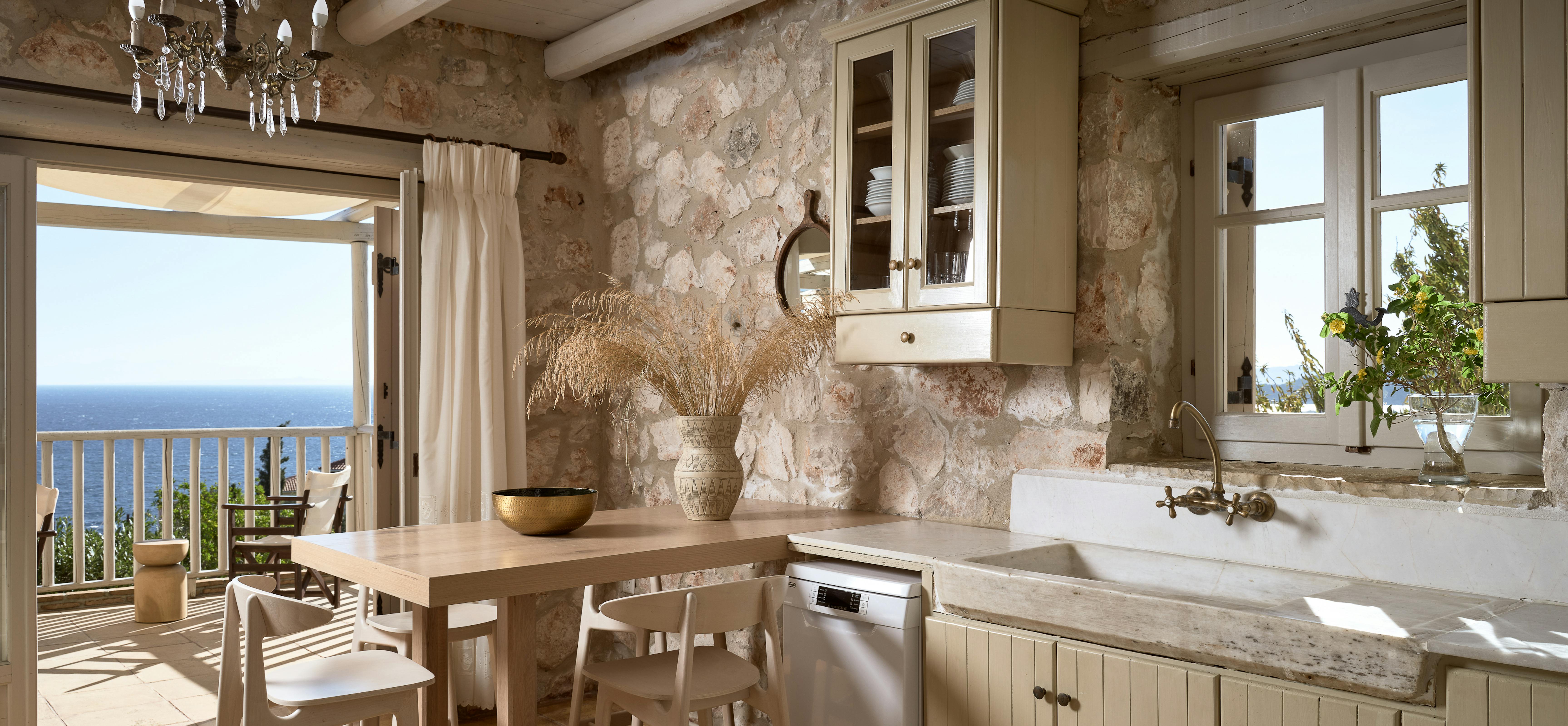 Kitchen with limestone walls, wooden dining table, vintage chandelier, white cabinetry with marble sink, and doors to sea-view balcony.