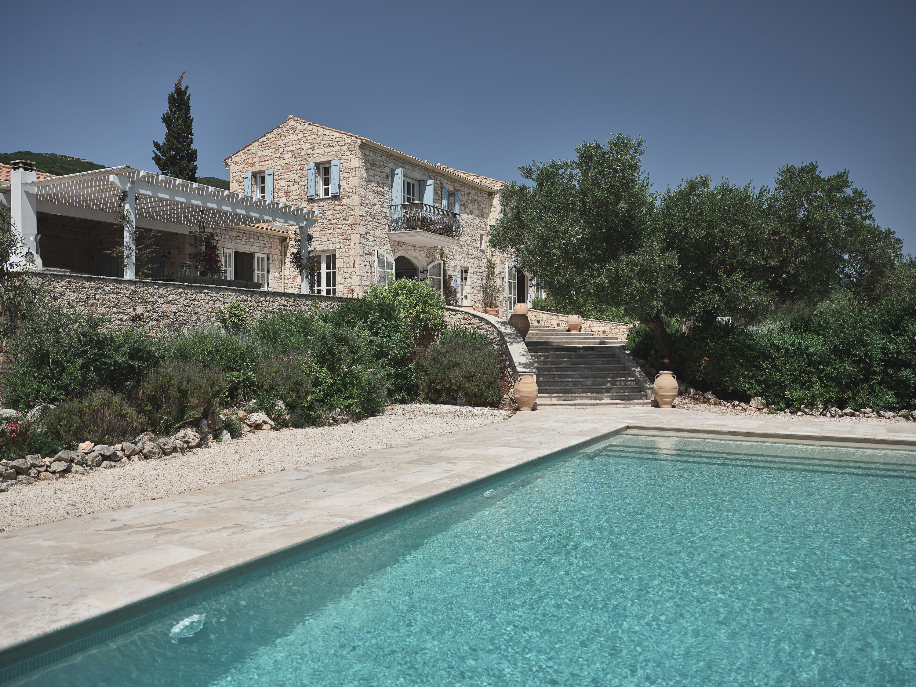 Hillside stone villa with pool overlooking the sea, surrounded by olive trees and Mediterranean landscaping.