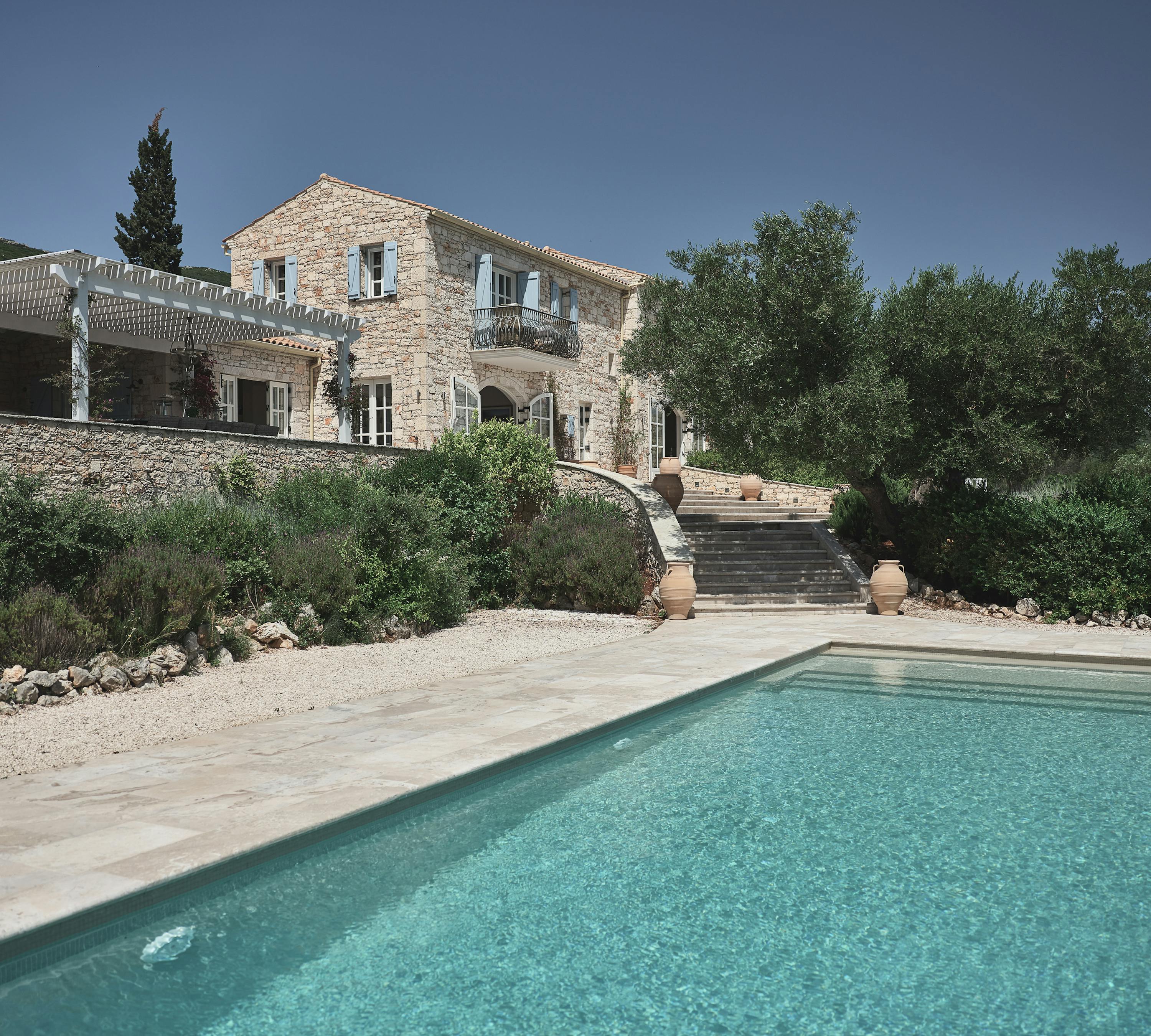 Hillside stone villa with pool overlooking the sea, surrounded by olive trees and Mediterranean landscaping.