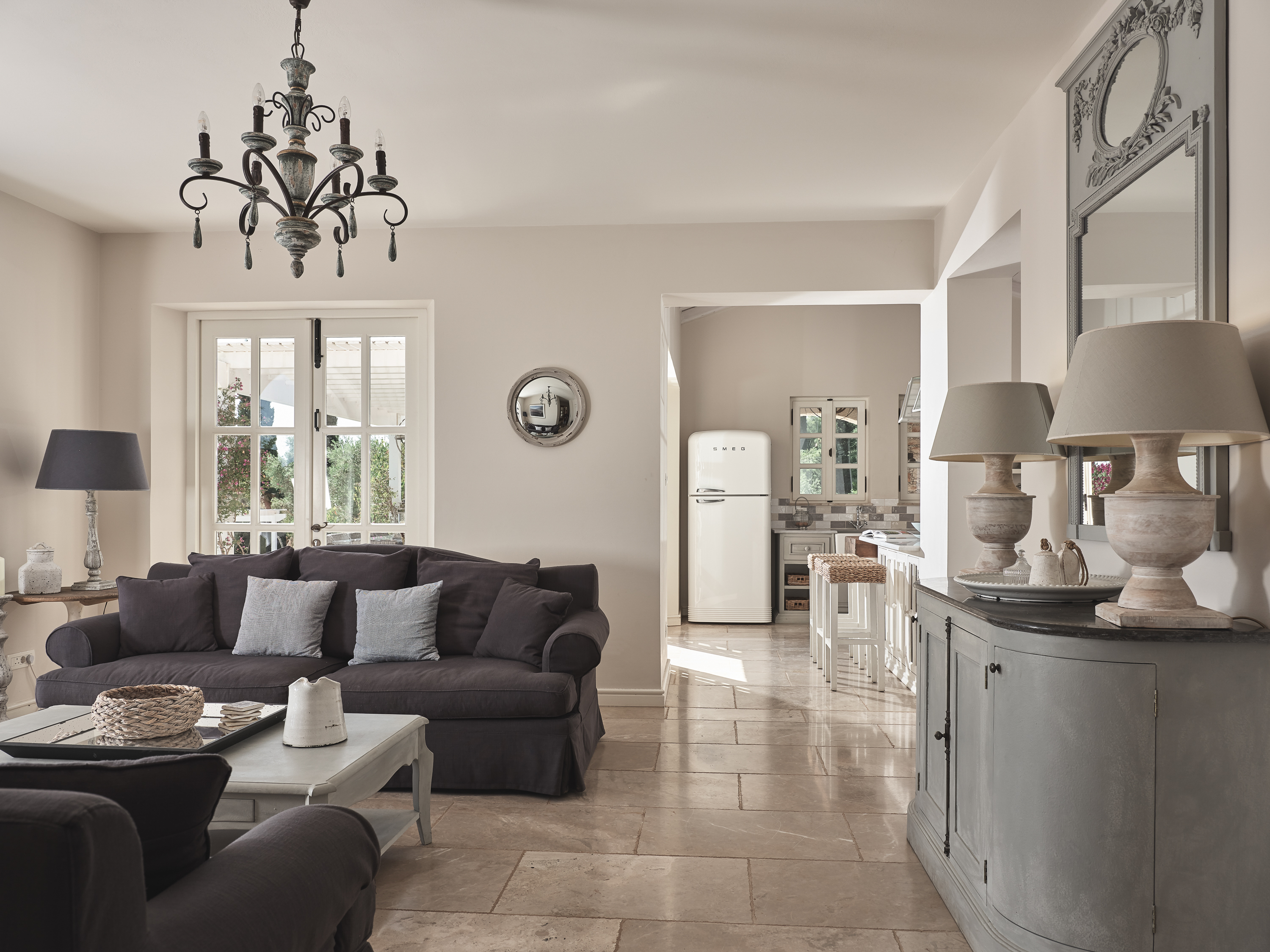 Open-plan living area with chandelier and dark sofas, connecting to bright farmhouse kitchen through white doorway.
