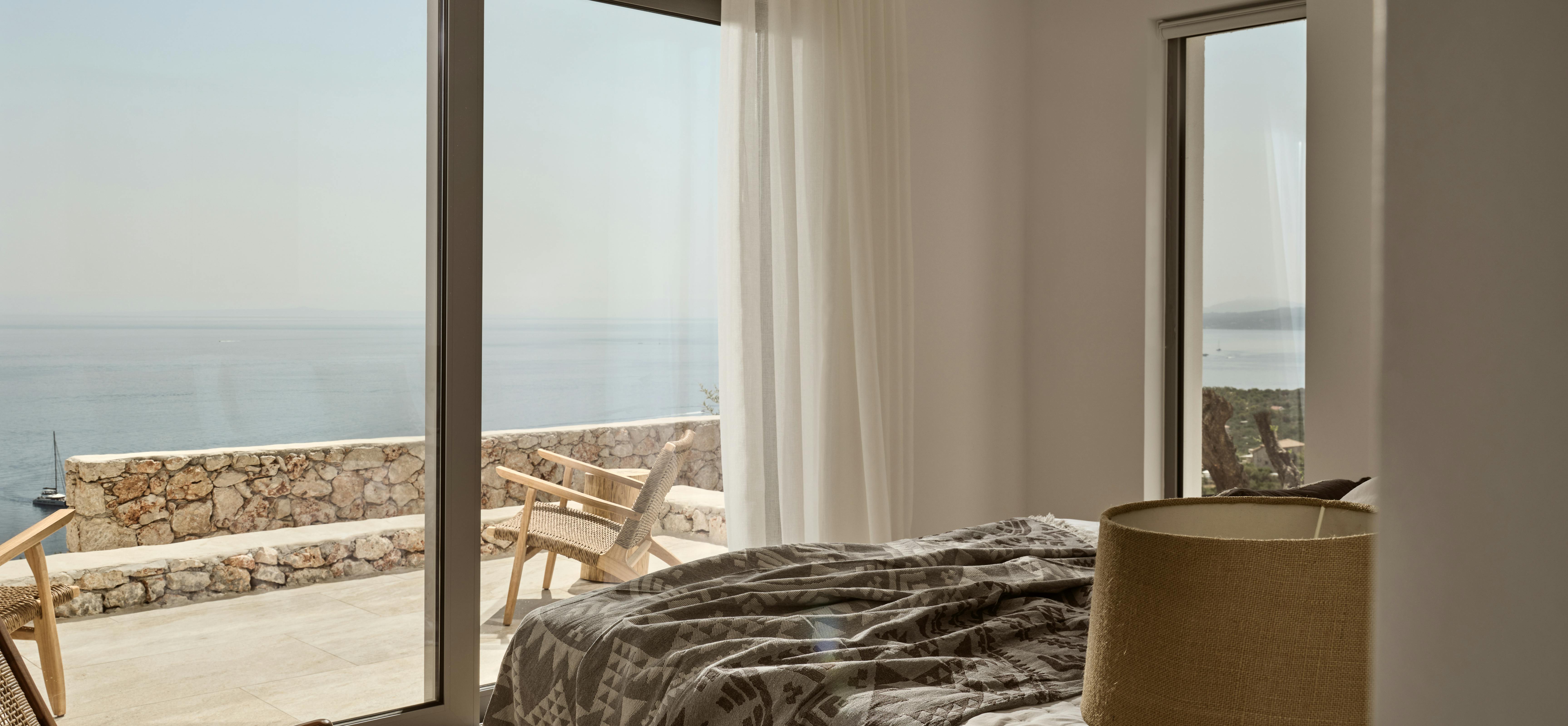 Bedroom with sliding glass doors opening to stone-walled terrace overlooking sea. Patterned throw blanket and woven chair visible on sun-drenched balcony.