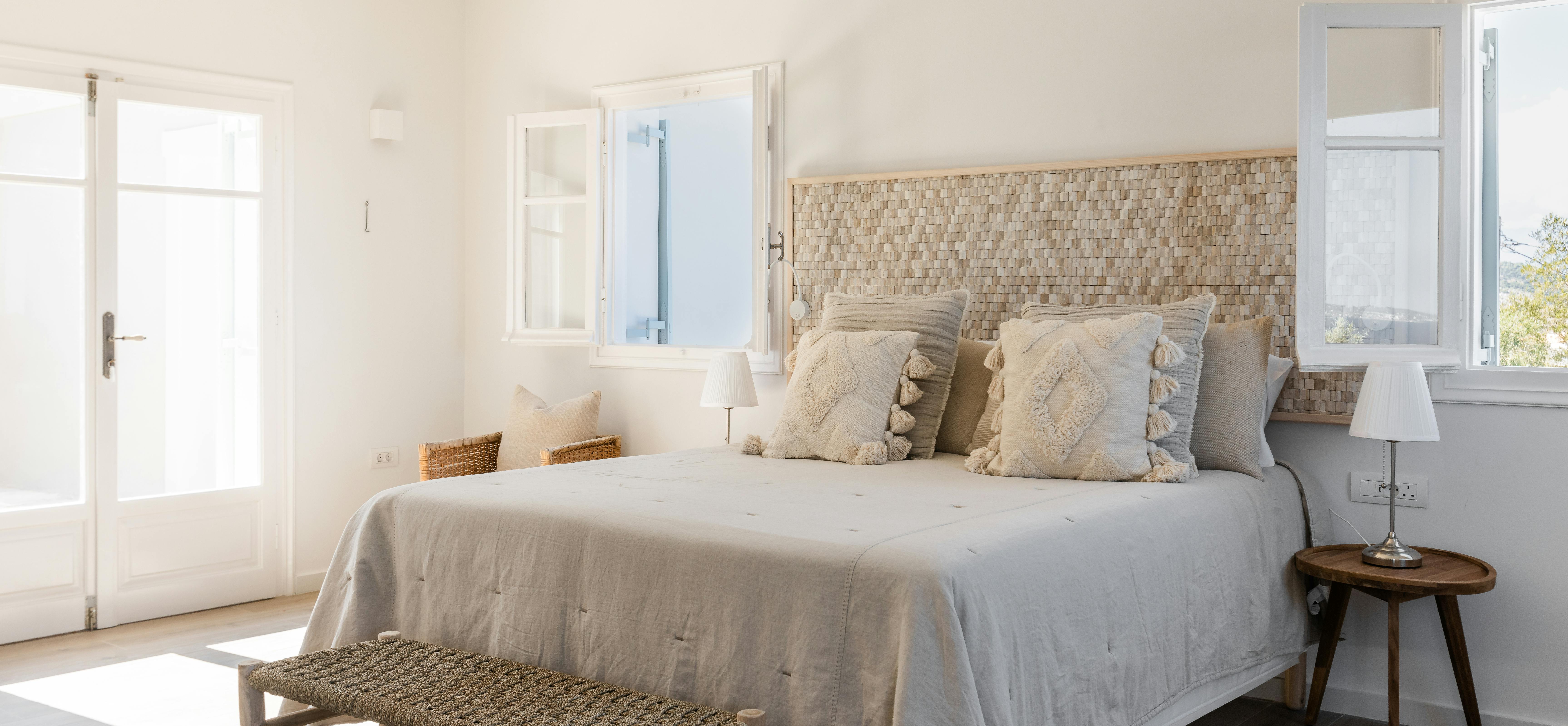 Minimalist white bedroom with natural woven headboard, neutral linens with tassel pillows, wooden bench at foot of bed, and French doors to terrace.