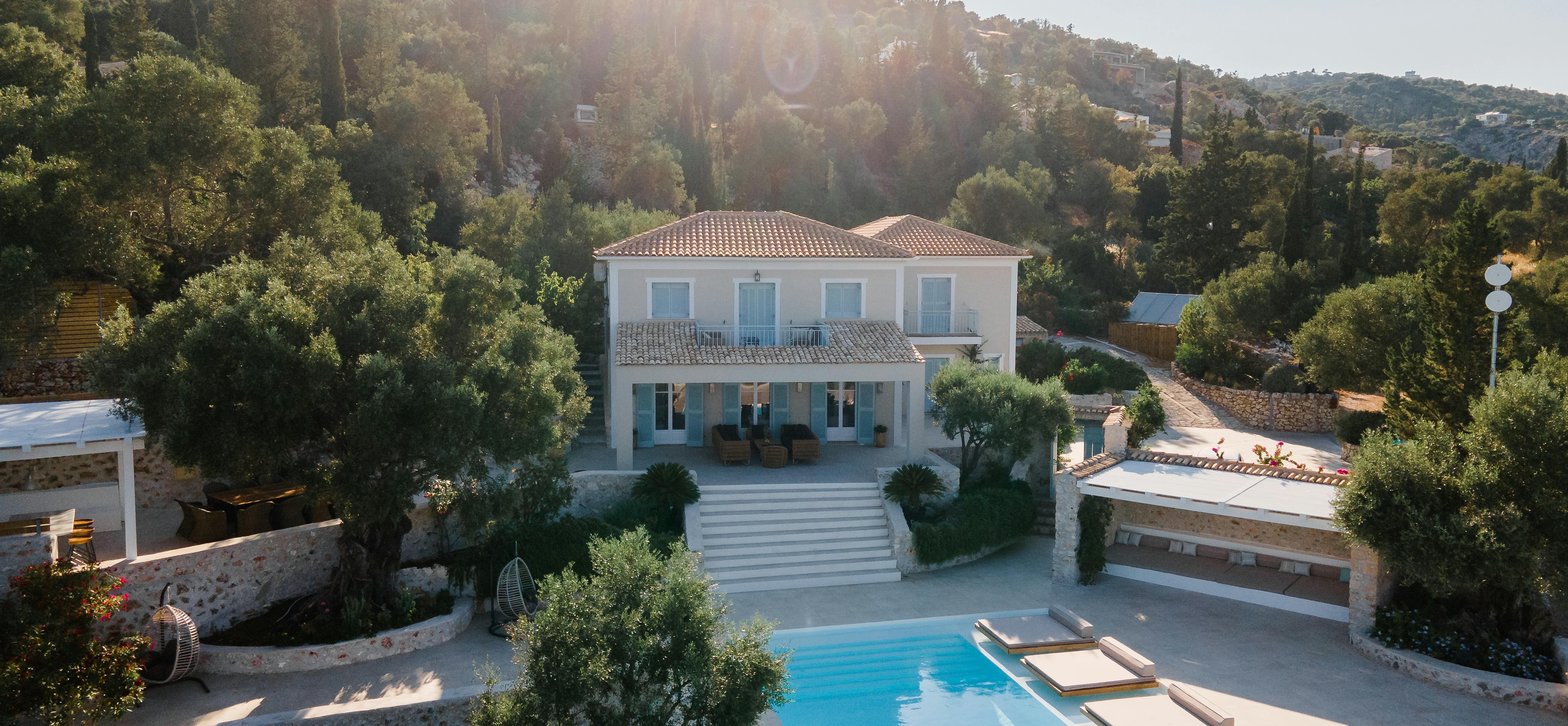 Aerial view of white Mediterranean villa with terracotta roof, infinity pool with loungers, stone terraces, and hillside setting with cypress trees.