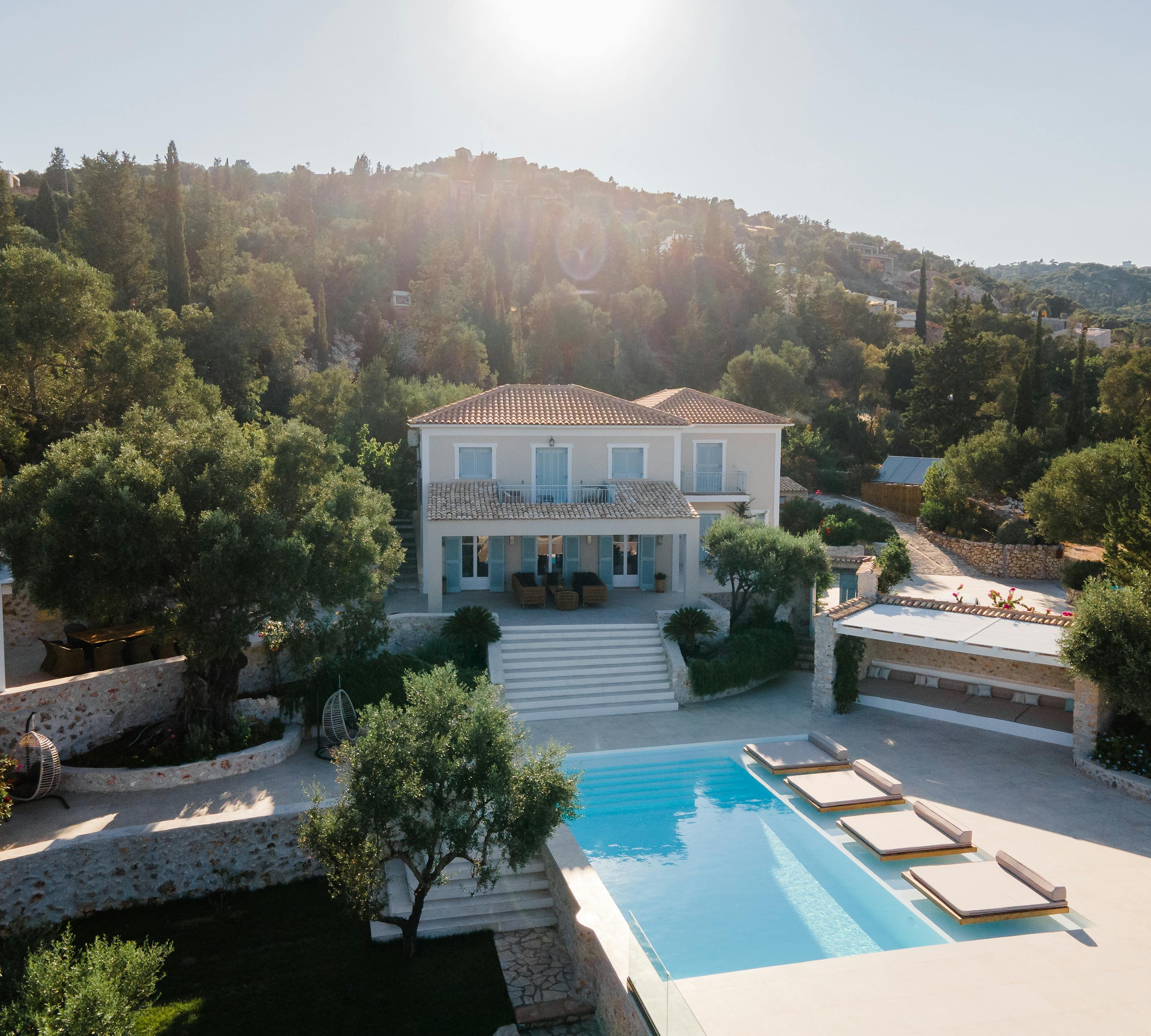 Aerial view of white Mediterranean villa with terracotta roof, infinity pool with loungers, stone terraces, and hillside setting with cypress trees.