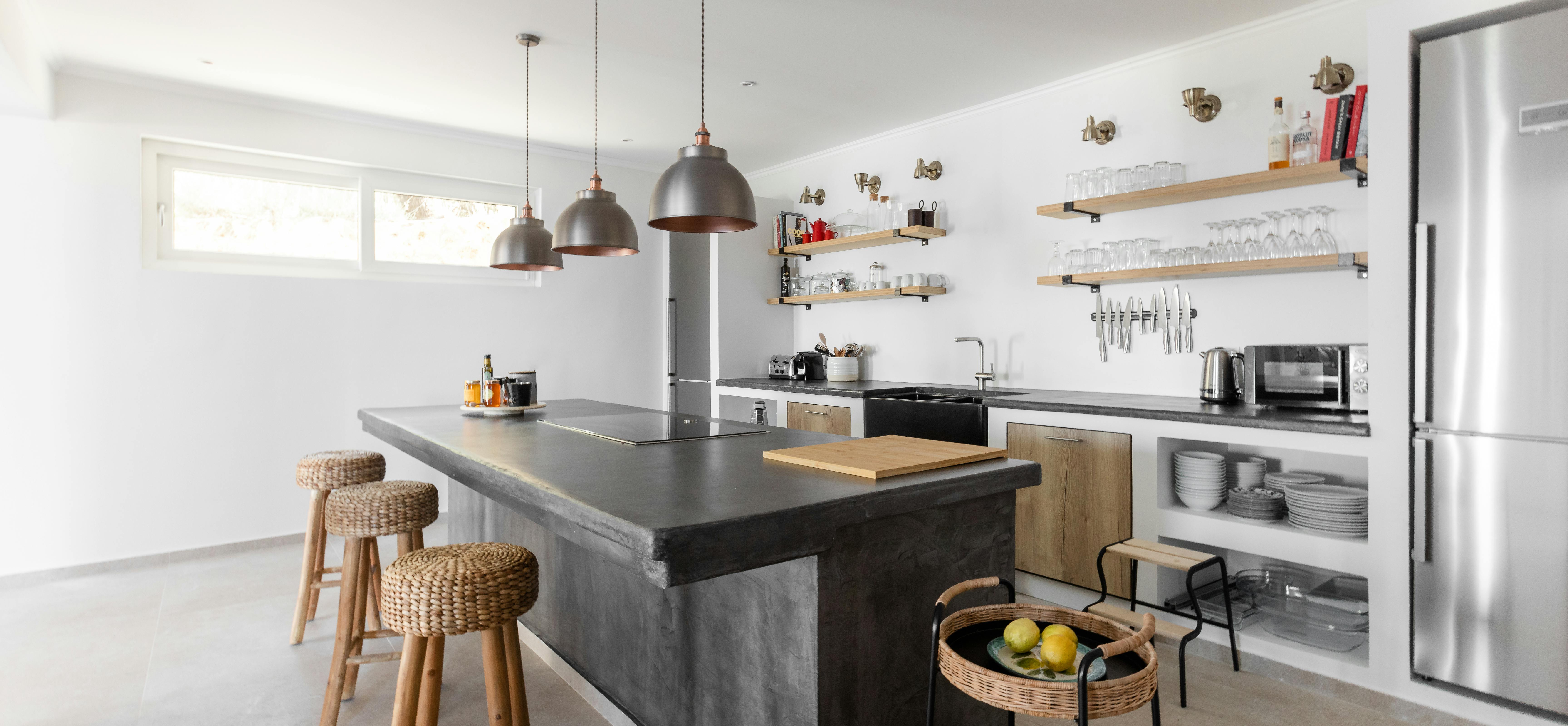 Contemporary kitchen with dark concrete island, woven bar stools, industrial pendant lights, and open wood shelving displaying glassware.