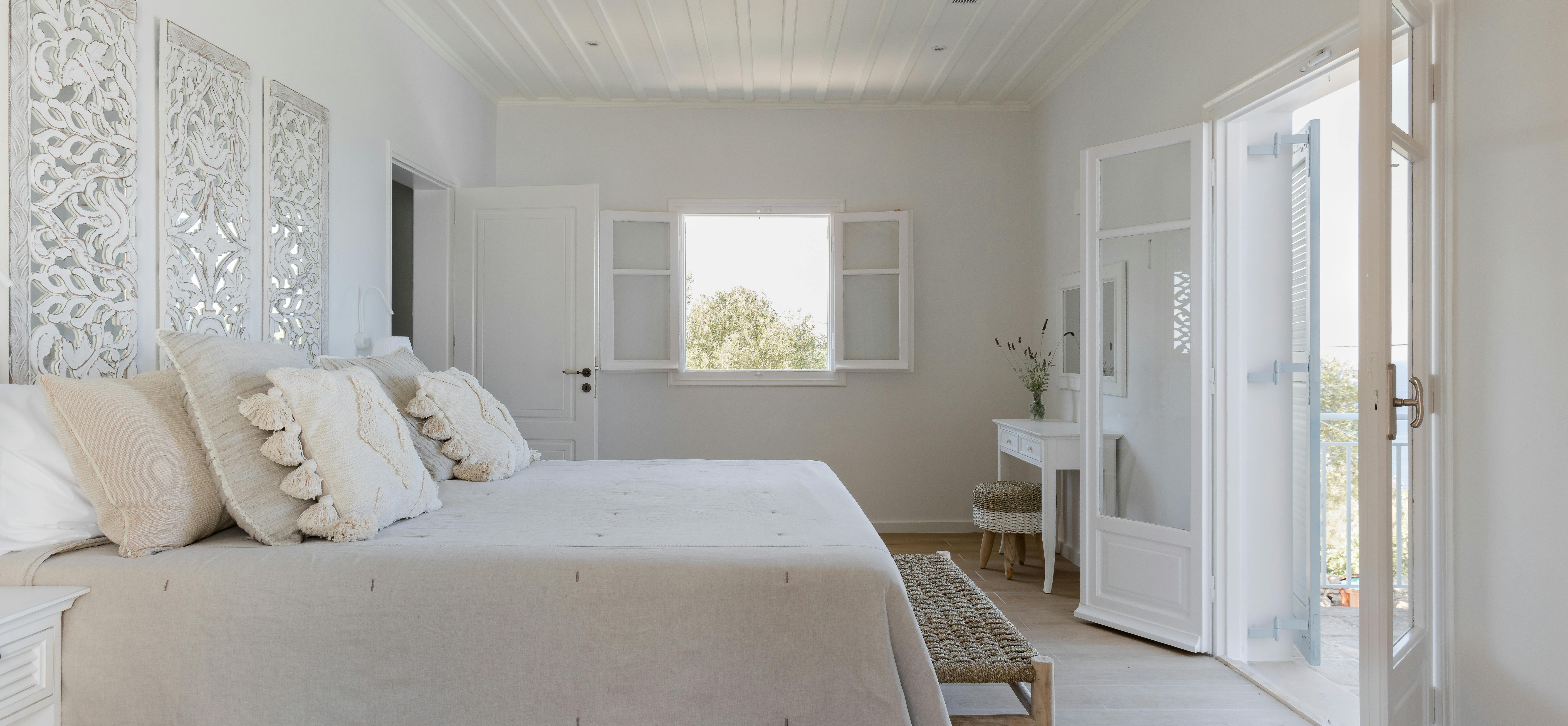 All-white master bedroom with carved decorative panels above bed, bench at foot, vanity area, and French doors opening to terrace.