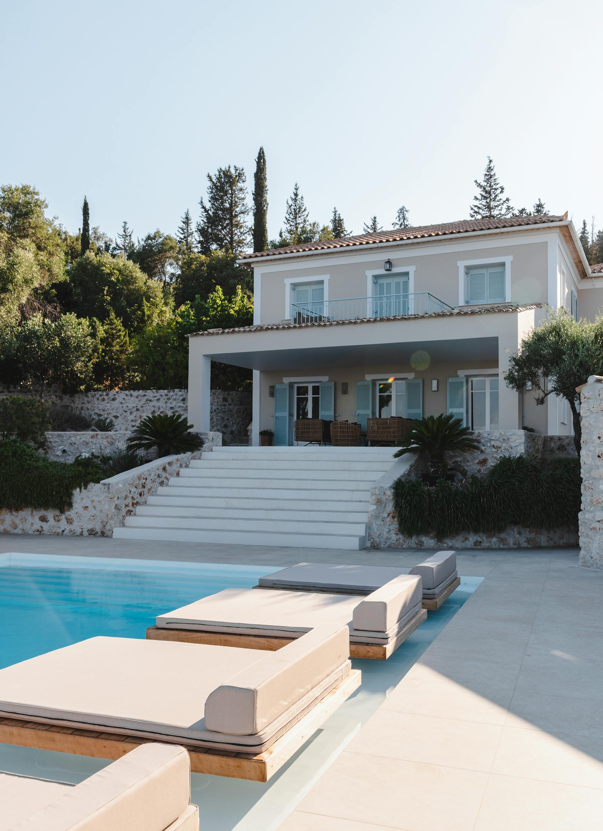 Pool deck view showing two-story white villa with blue shutters, grand staircase, stone walls, and modern loungers beside infinity pool.