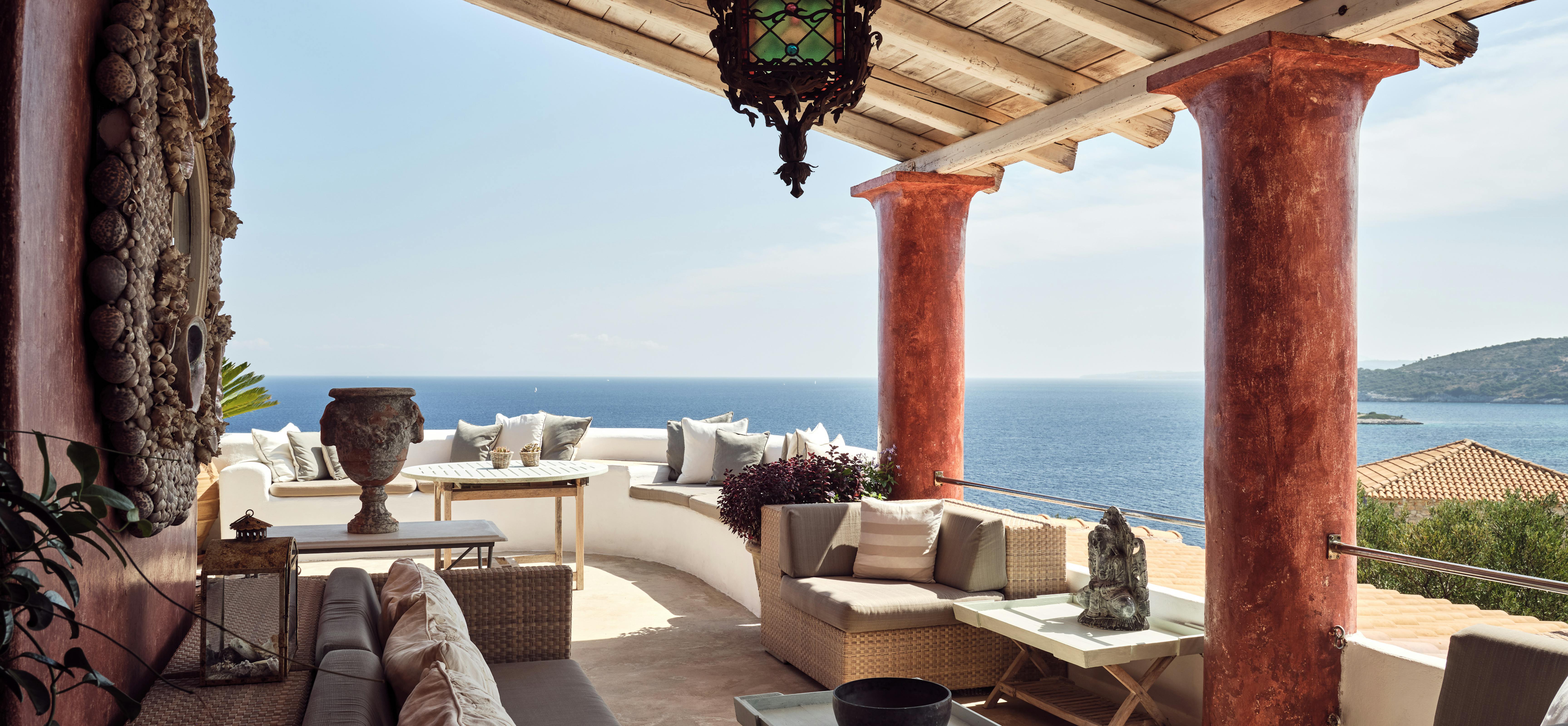 Expansive covered terrace with weathered terracotta columns, ornate lantern, curved white sofas, wicker furniture, and panoramic Mediterranean Sea views.