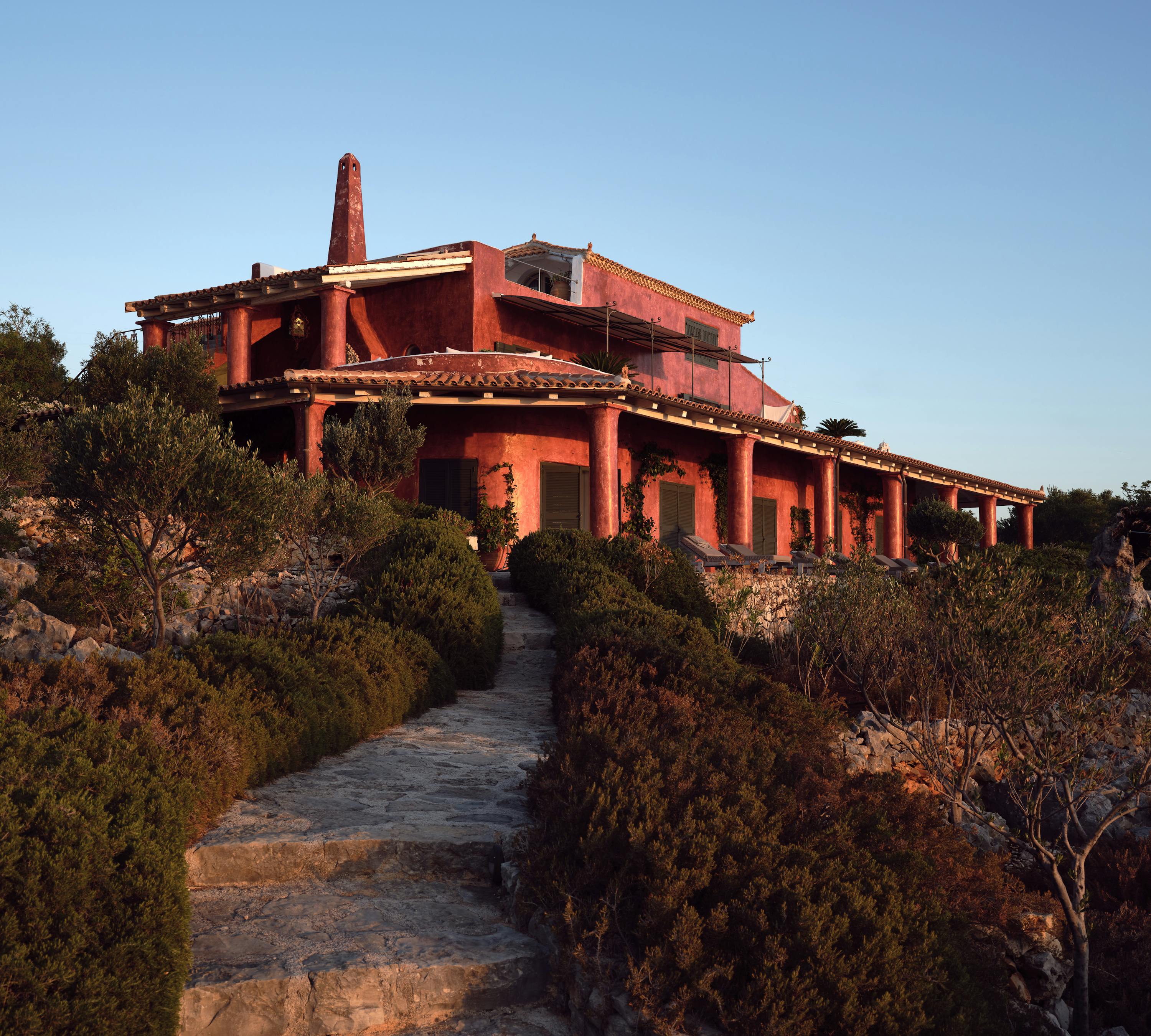 Dramatic terracotta-coloured Mediterranean villa at dusk perched on hillside with multiple levels, pergolas, and winding stone path through native vegetation.