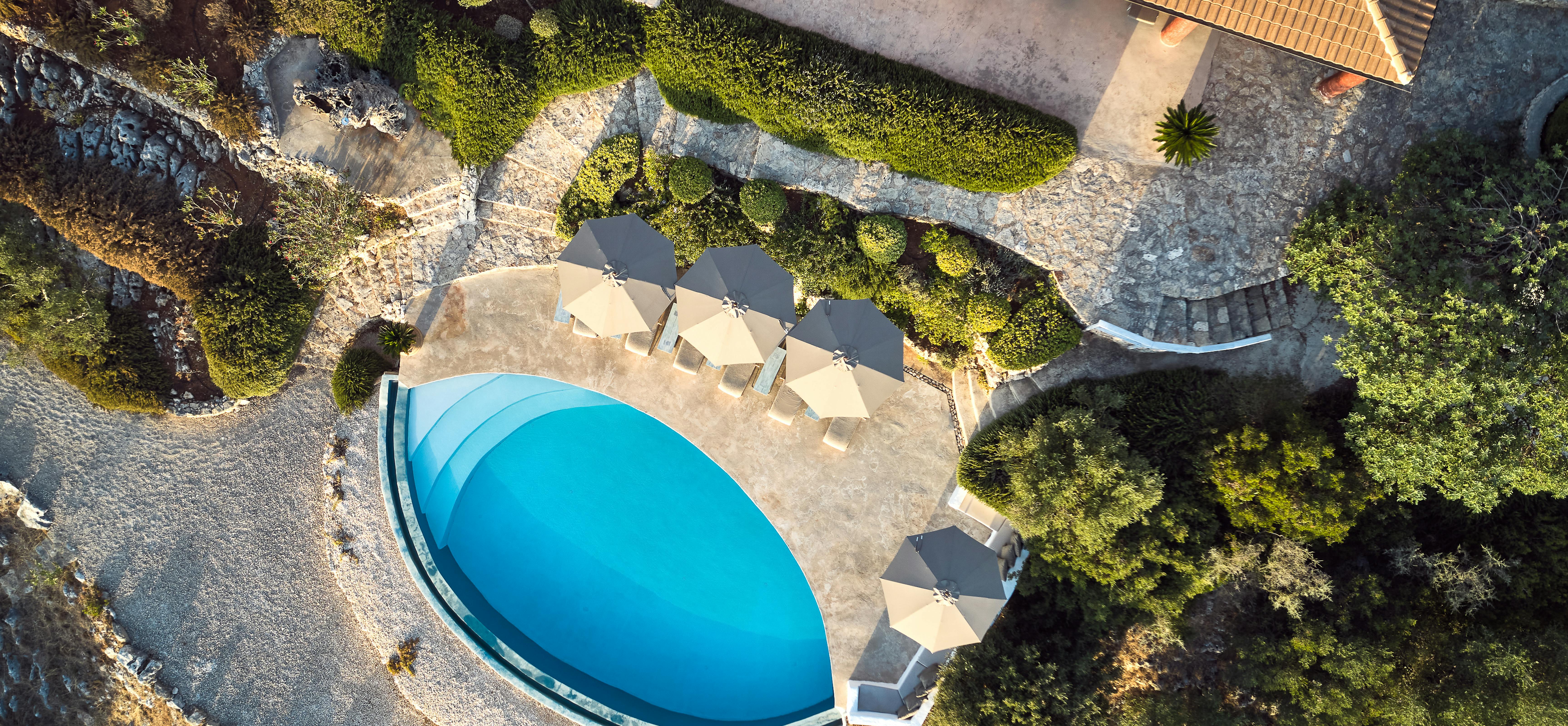 Aerial view of curved infinity pool with beige umbrellas, stone terrace, natural landscaping, and terracotta-roofed villa visible above.
