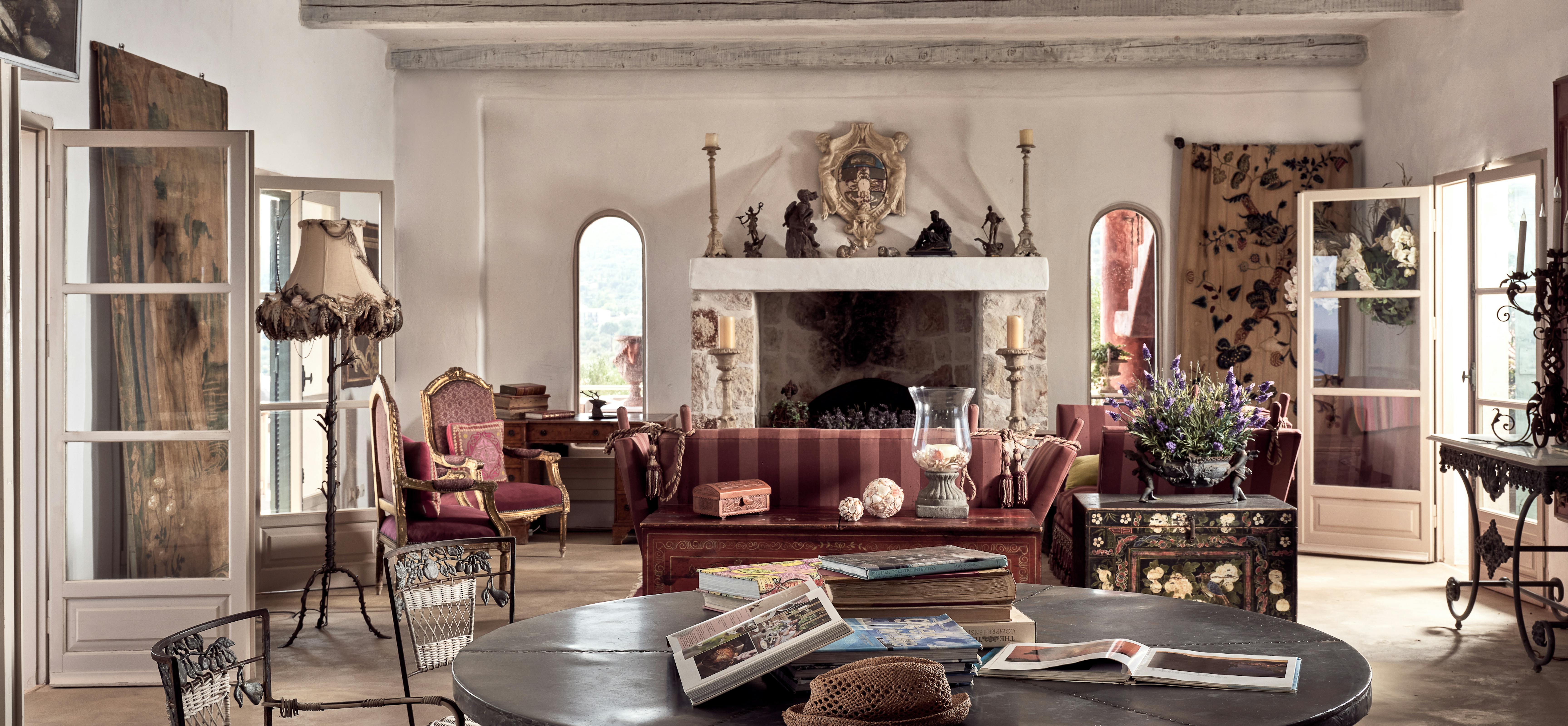 Grand living room with exposed beams, ornate stone fireplace, burgundy velvet furniture, large round table with wrought iron chairs, and arched doorways.