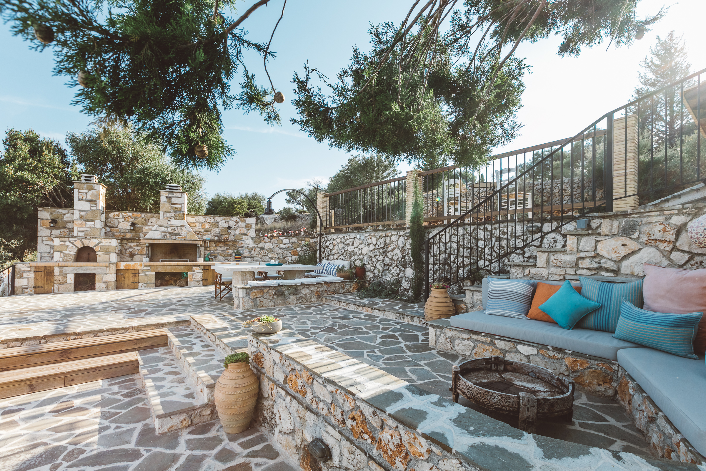 Multi-level stone terrace with irregular flagstone paving, built-in turquoise cushioned bench seating, outdoor fireplace and barbecue, large urns, and pine tree canopy.