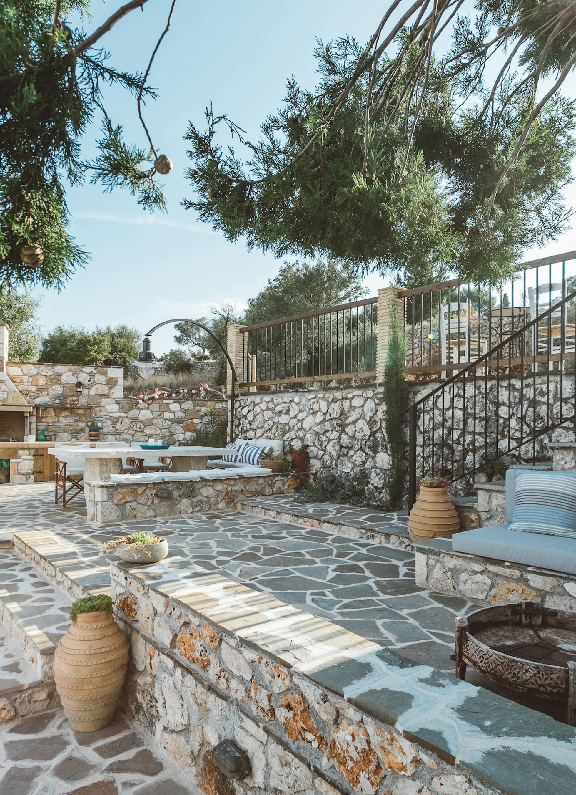 Multi-level stone terrace with irregular flagstone paving, built-in turquoise cushioned bench seating, outdoor fireplace and barbecue, large urns, and pine tree canopy.