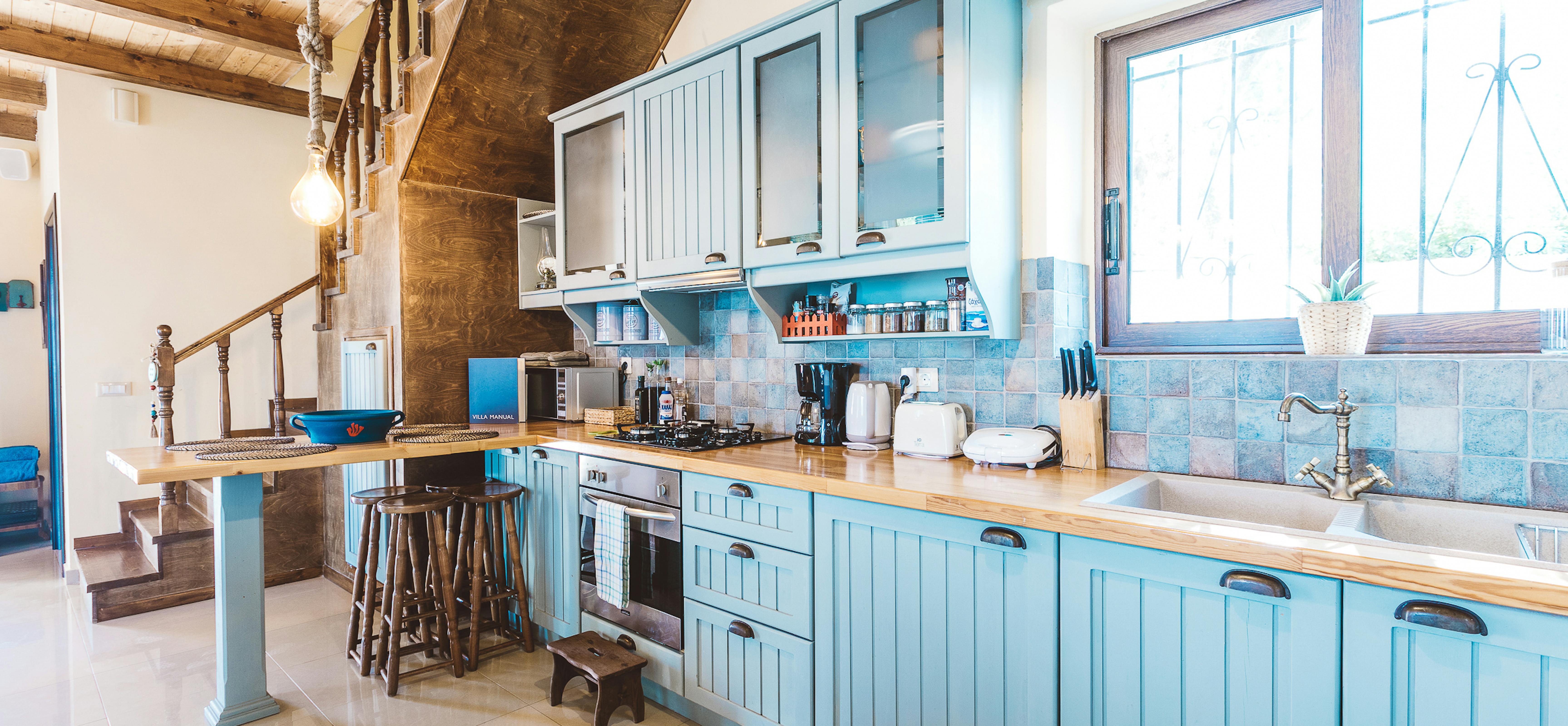 Cottage-style kitchen with powder blue headboard cabinetry, butcher block counters, iridescent blue tile backsplash, breakfast bar, and exposed wood ceiling with stairs.