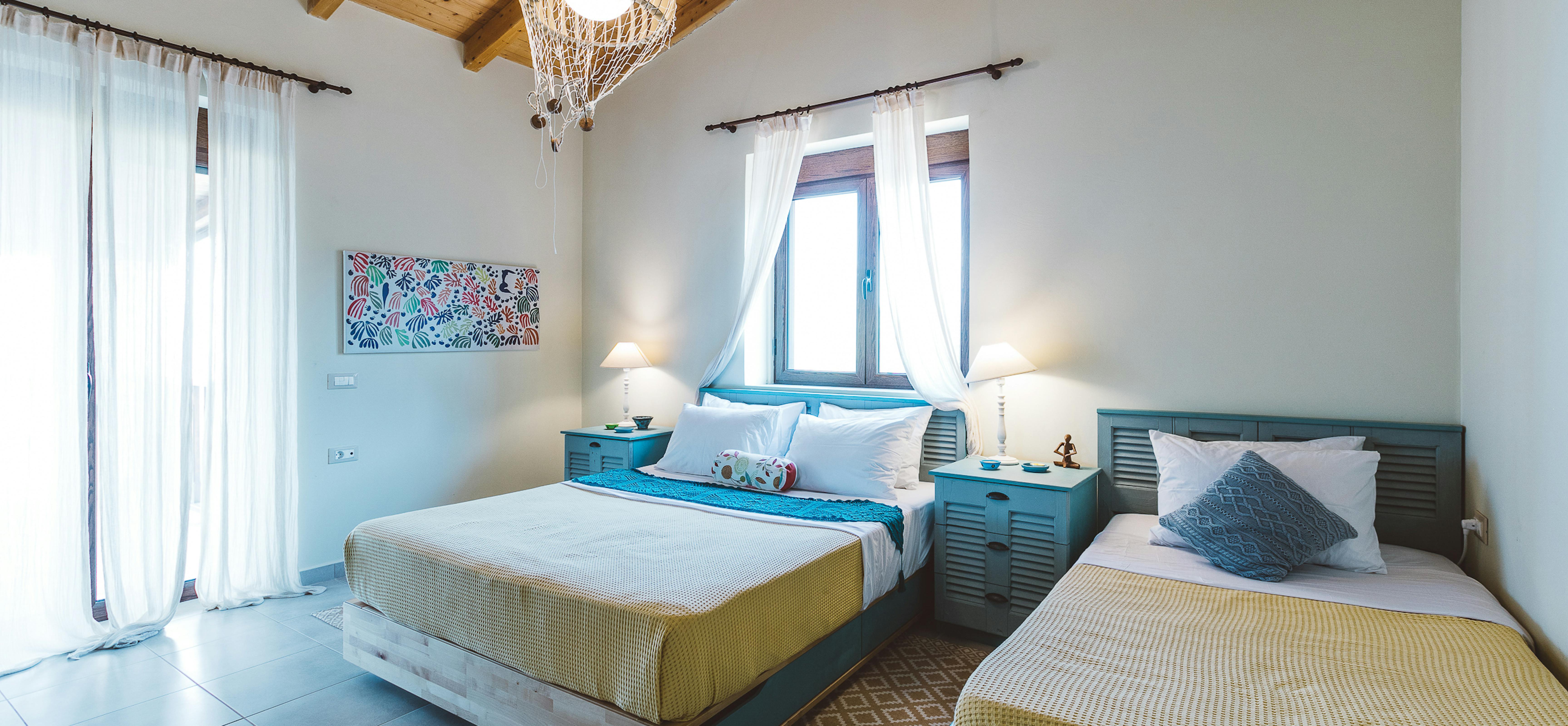 Twin bedroom with vaulted wood ceiling, turquoise louvered headboards, colourful artwork, wicker pendant light, white curtains, and yellow waffle-weave bedding.
