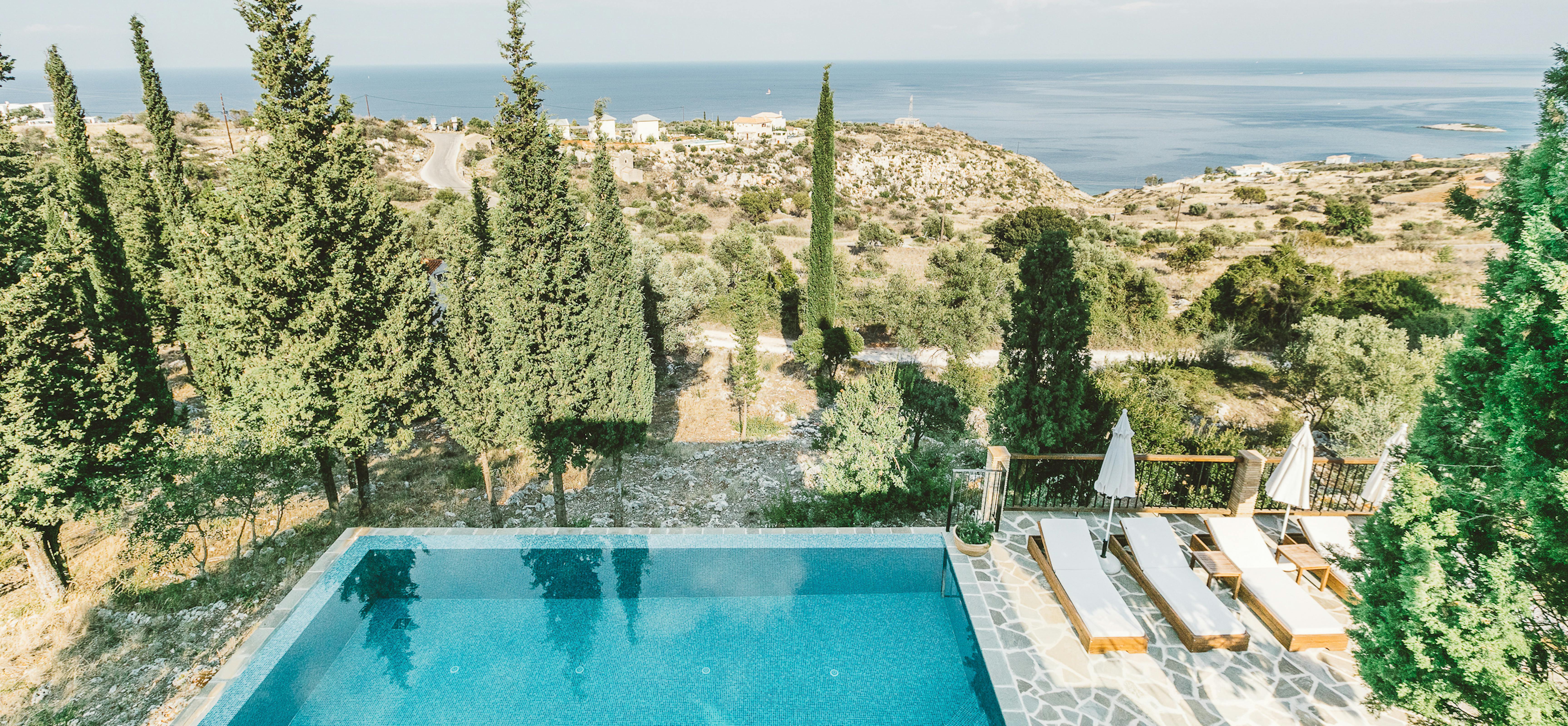 Elevated pool deck with wooden loungers, white umbrellas, irregular stone paving, cypress and pine trees, and panoramic coastal and countryside views.