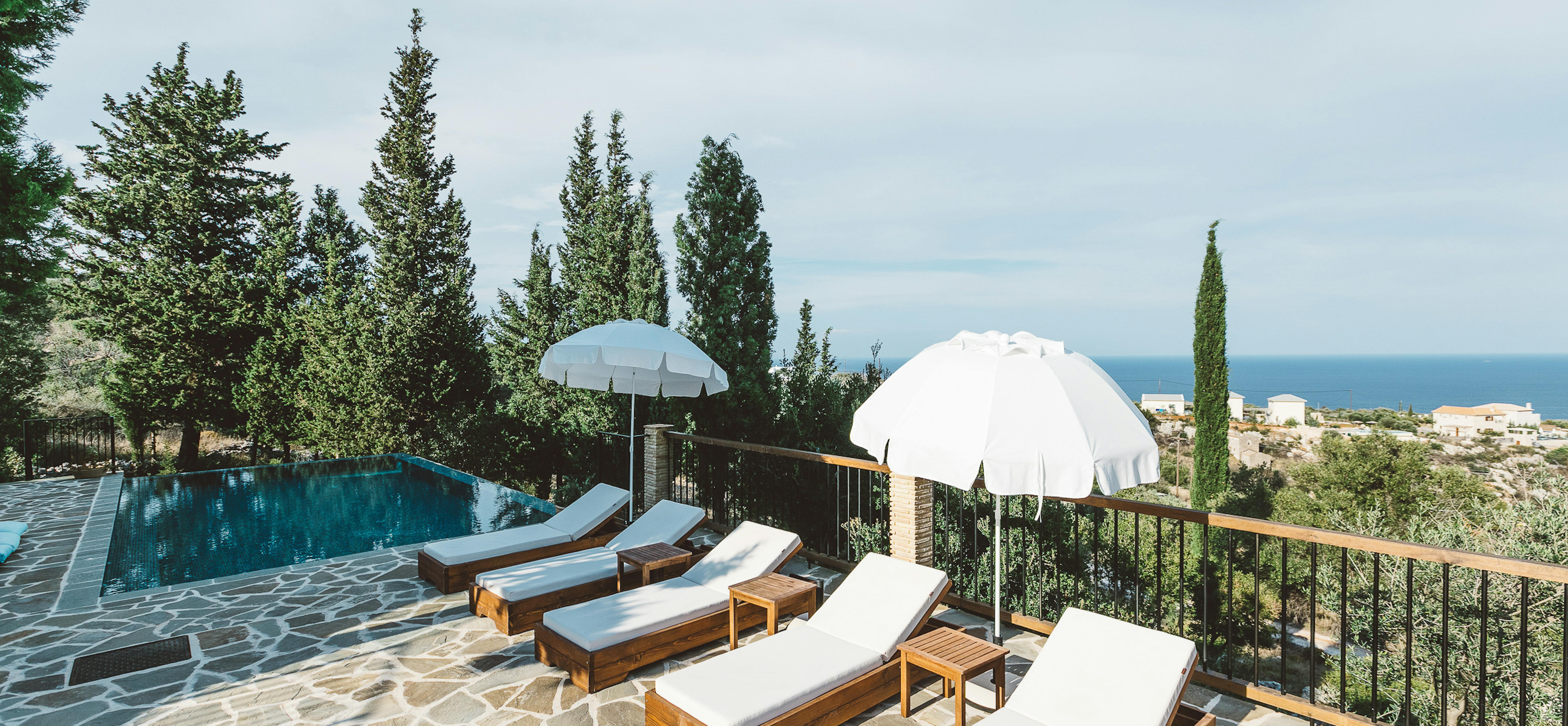 Pool terrace with wooden loungers, white umbrella, irregular stone paving, glass safety railing, cypress trees, and distant sea and town views.