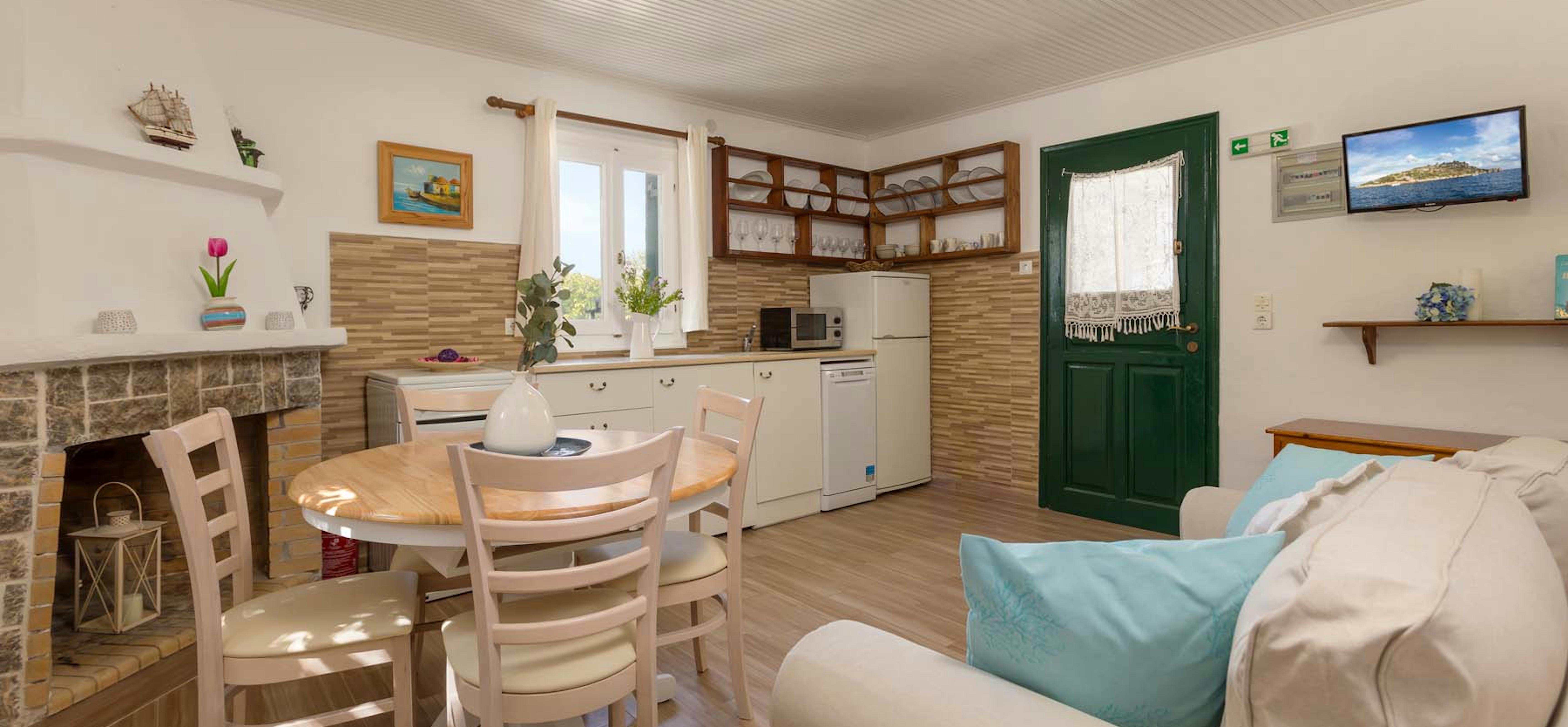 Bright open-plan vacation cottage interior with kitchen, dining area, white sofa with turquoise cushions, and green door.
