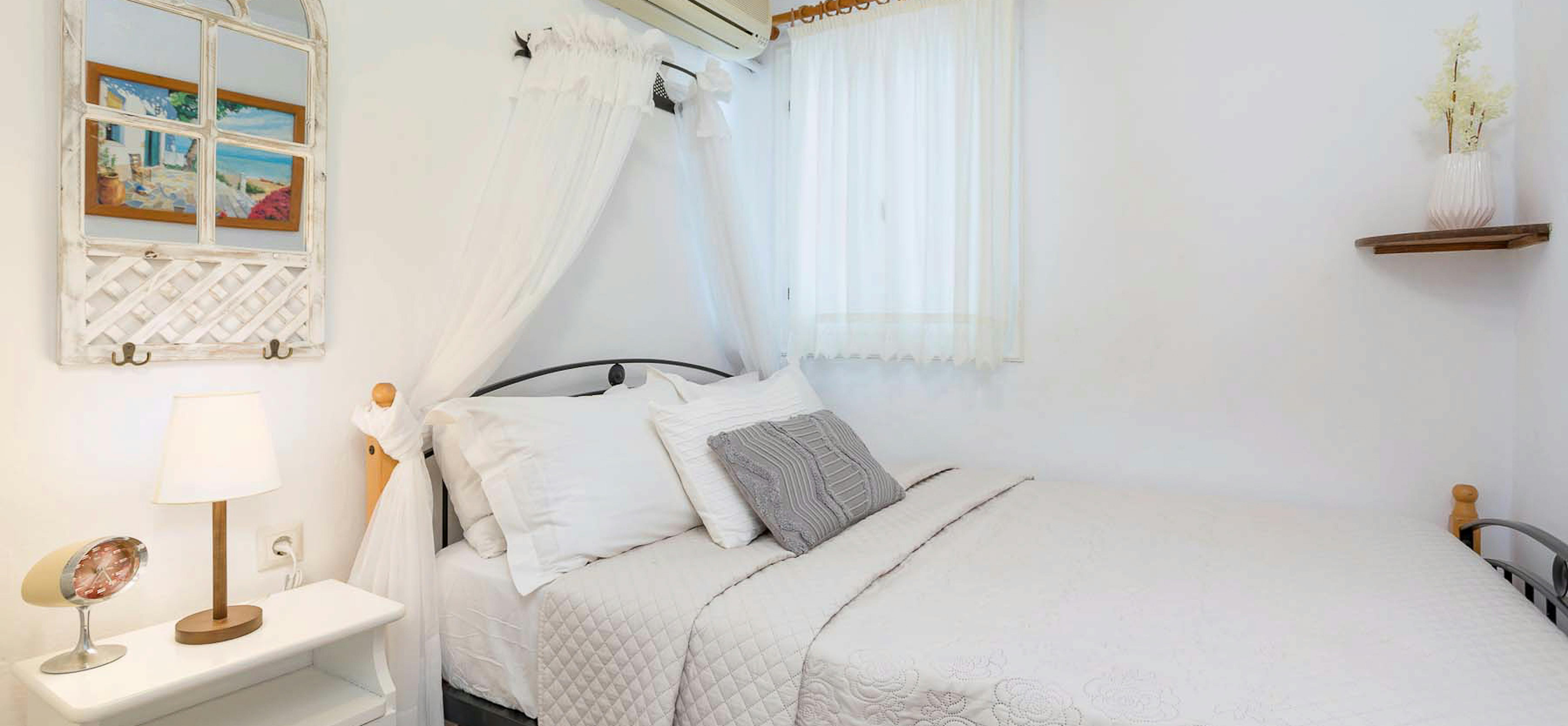 Bright bedroom with white bedding, mosquito net canopy, air conditioning, and decorative window mirror.