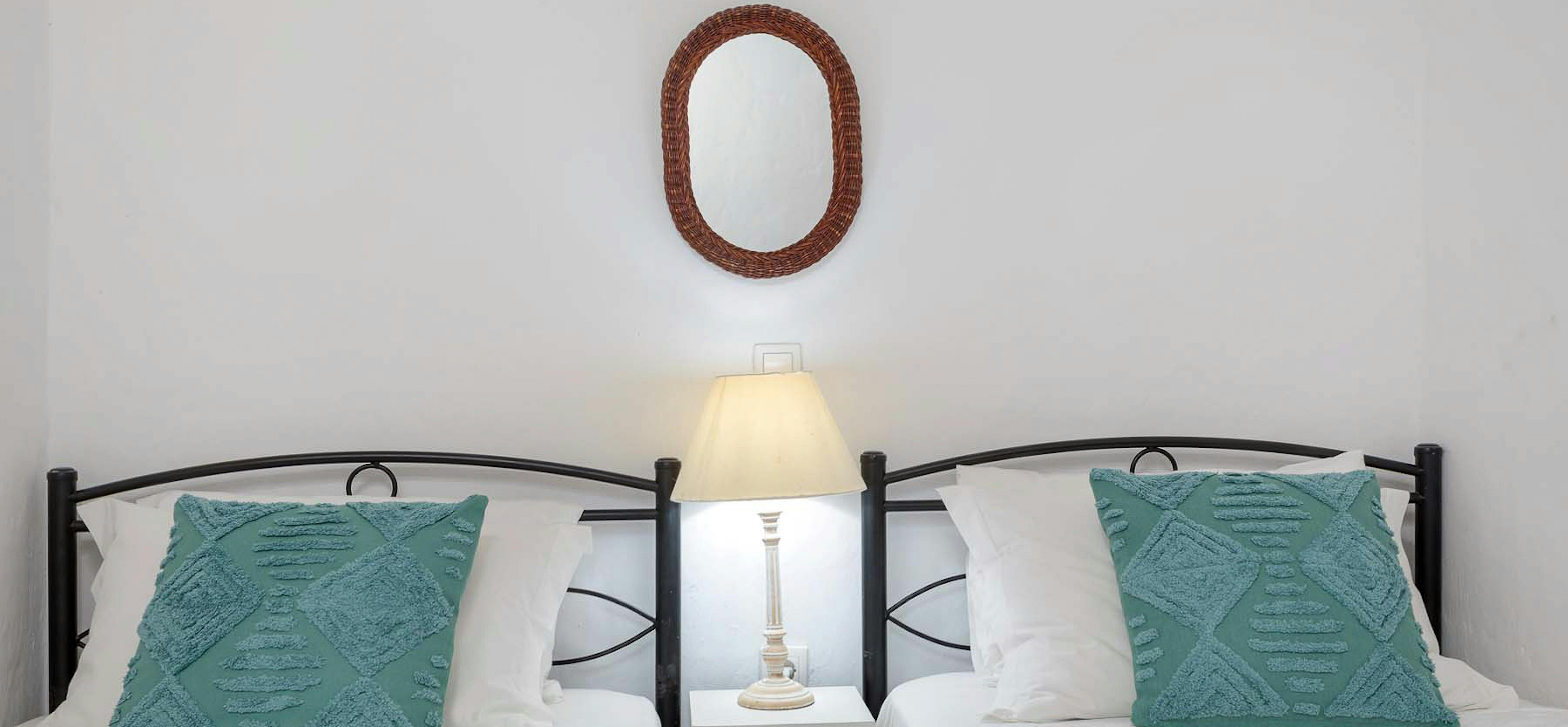 Twin bedroom with black metal bed frames, teal cushions, white nightstand, and woven mirror.