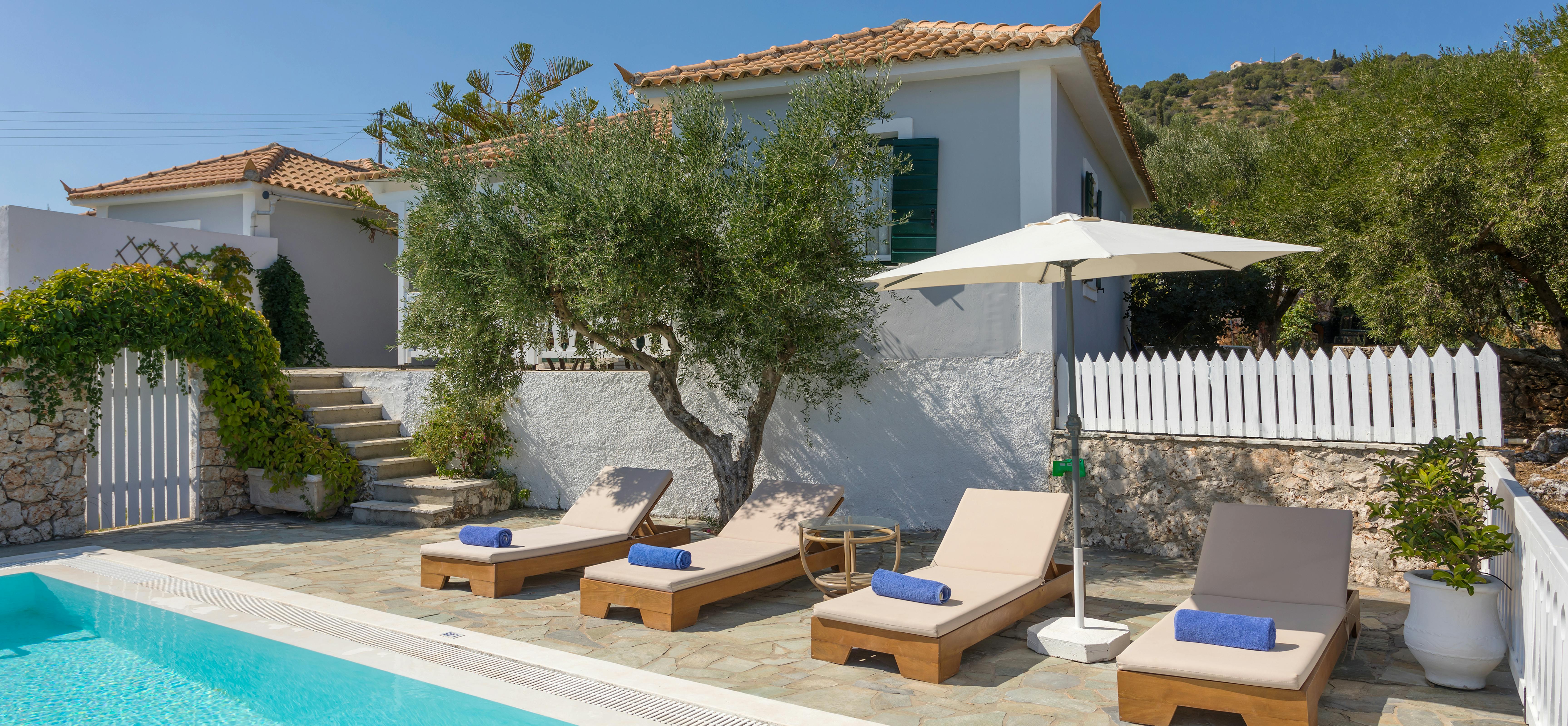 Villa pool terrace with four wooden loungers, white umbrella, olive tree, and white picket fencing.