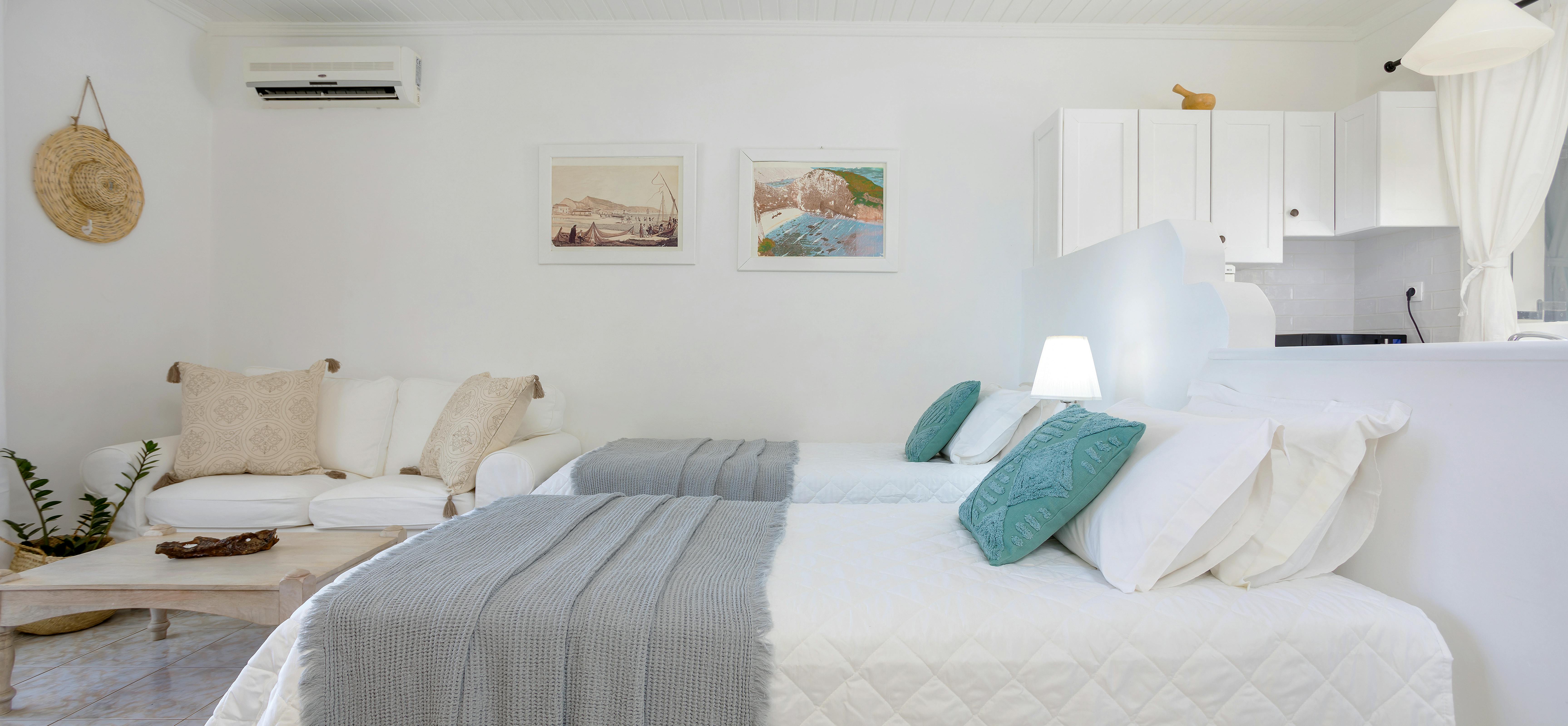 White studio bedroom with twin beds, grey throws, turquoise cushions, seating area, and kitchenette.