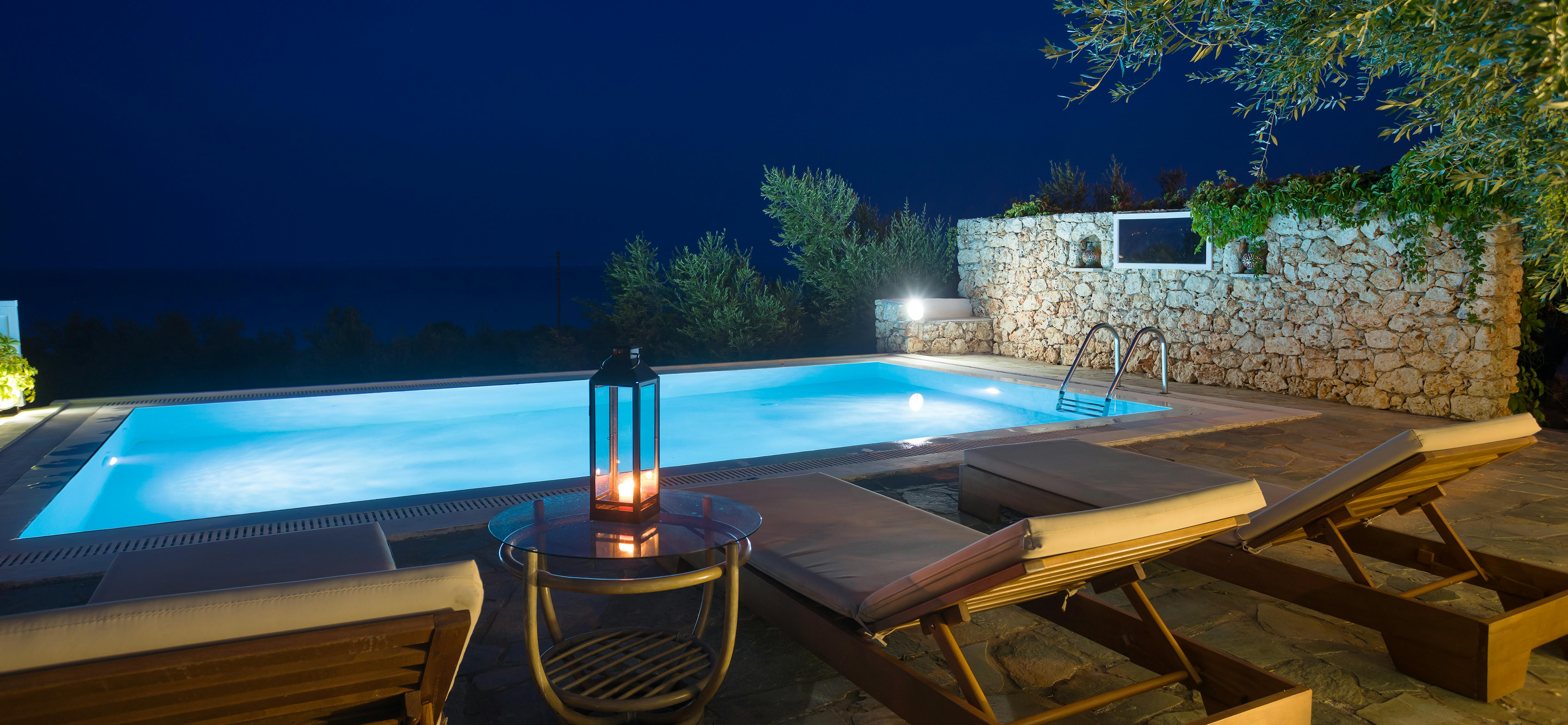 Night view of illuminated swimming pool with wooden loungers, lantern, and stone wall backdrop.