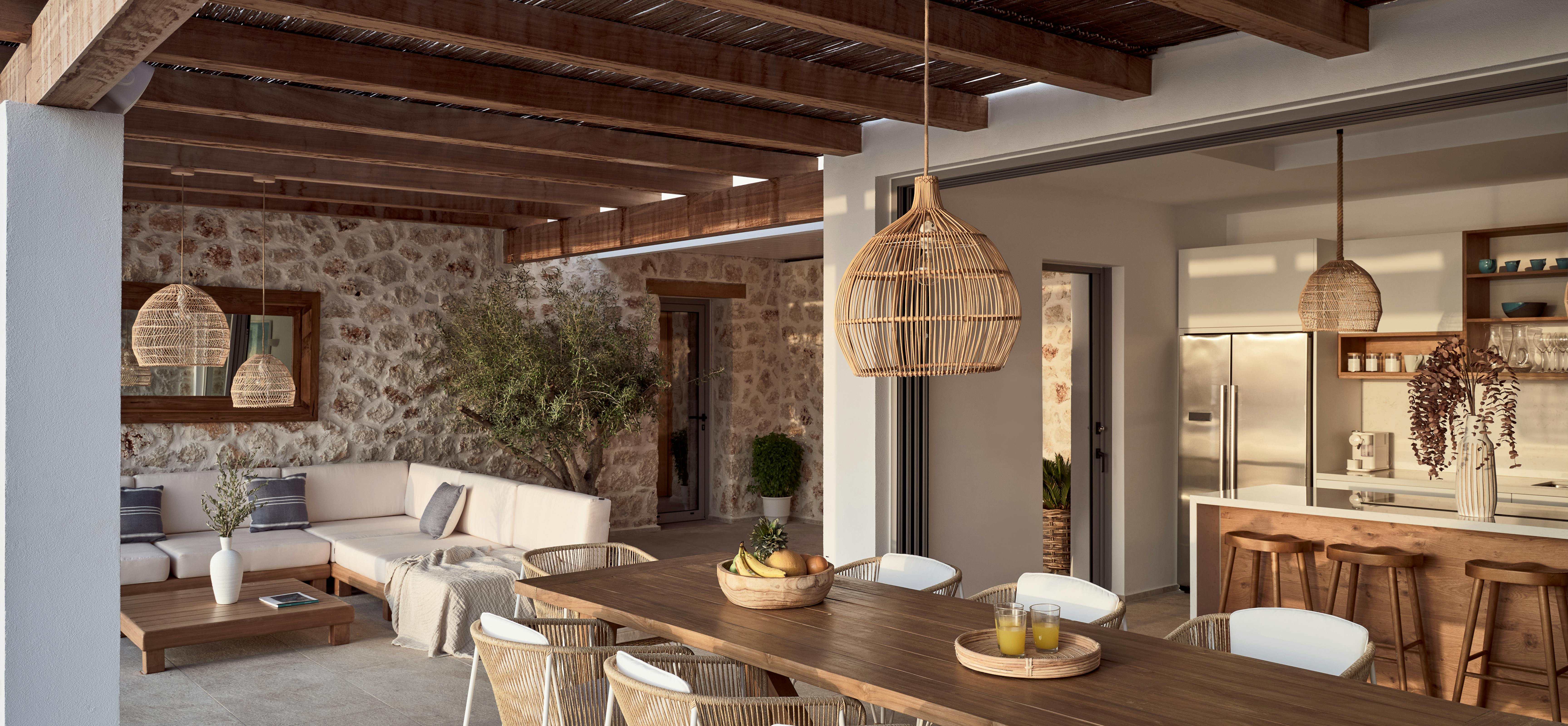 Open-air dining space with wooden table, wicker chairs, stone walls, and modern kitchen visible beyond. Exposed beam ceiling and woven pendant lights complement the Mediterranean aesthetic.