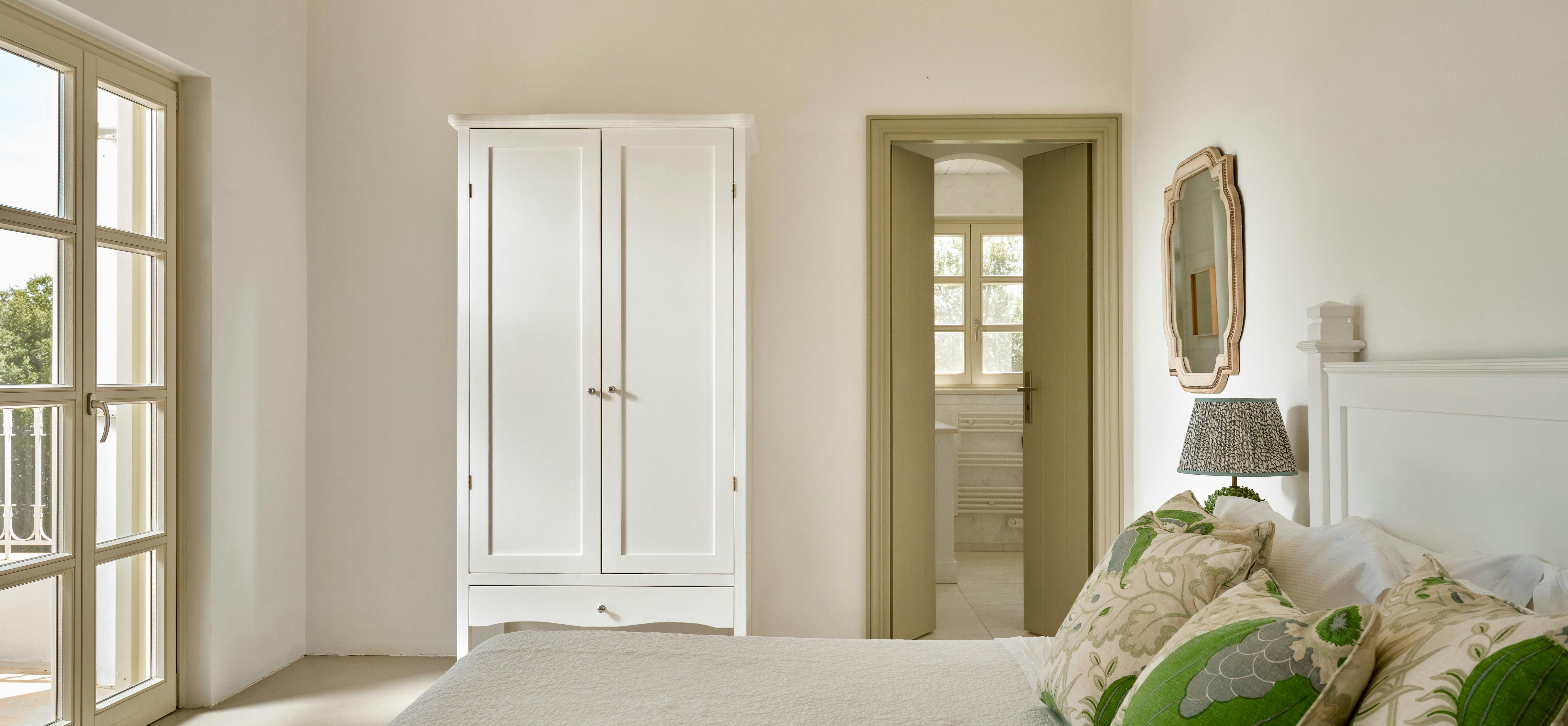 Bright bedroom suite with white armoire and green botanical throw pillows. Large windows flood space with natural light, overlooking garden views.
