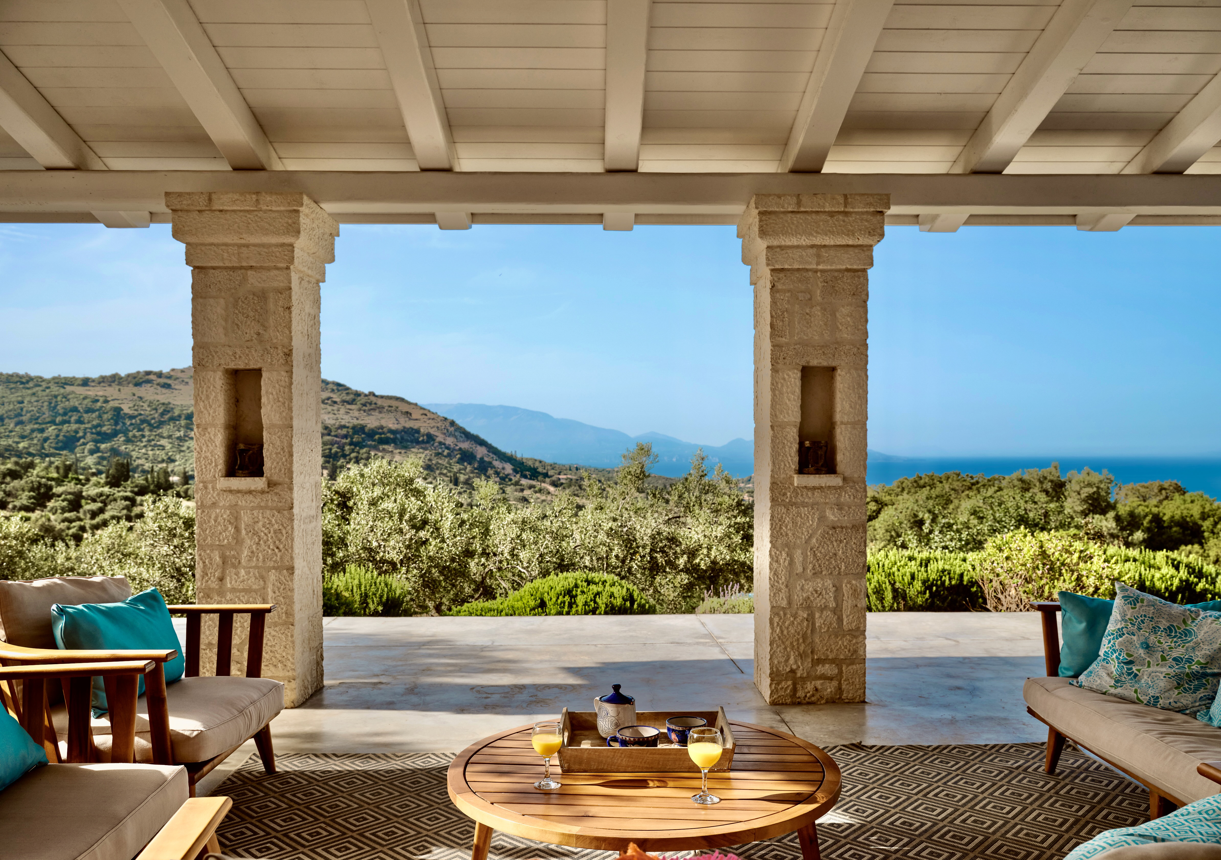 Covered terrace with stone columns and exposed beam ceiling framing panoramic views of olive groves and distant coastline. Teak lounge furniture with turquoise cushions arranged around round coffee table set for breakfast.