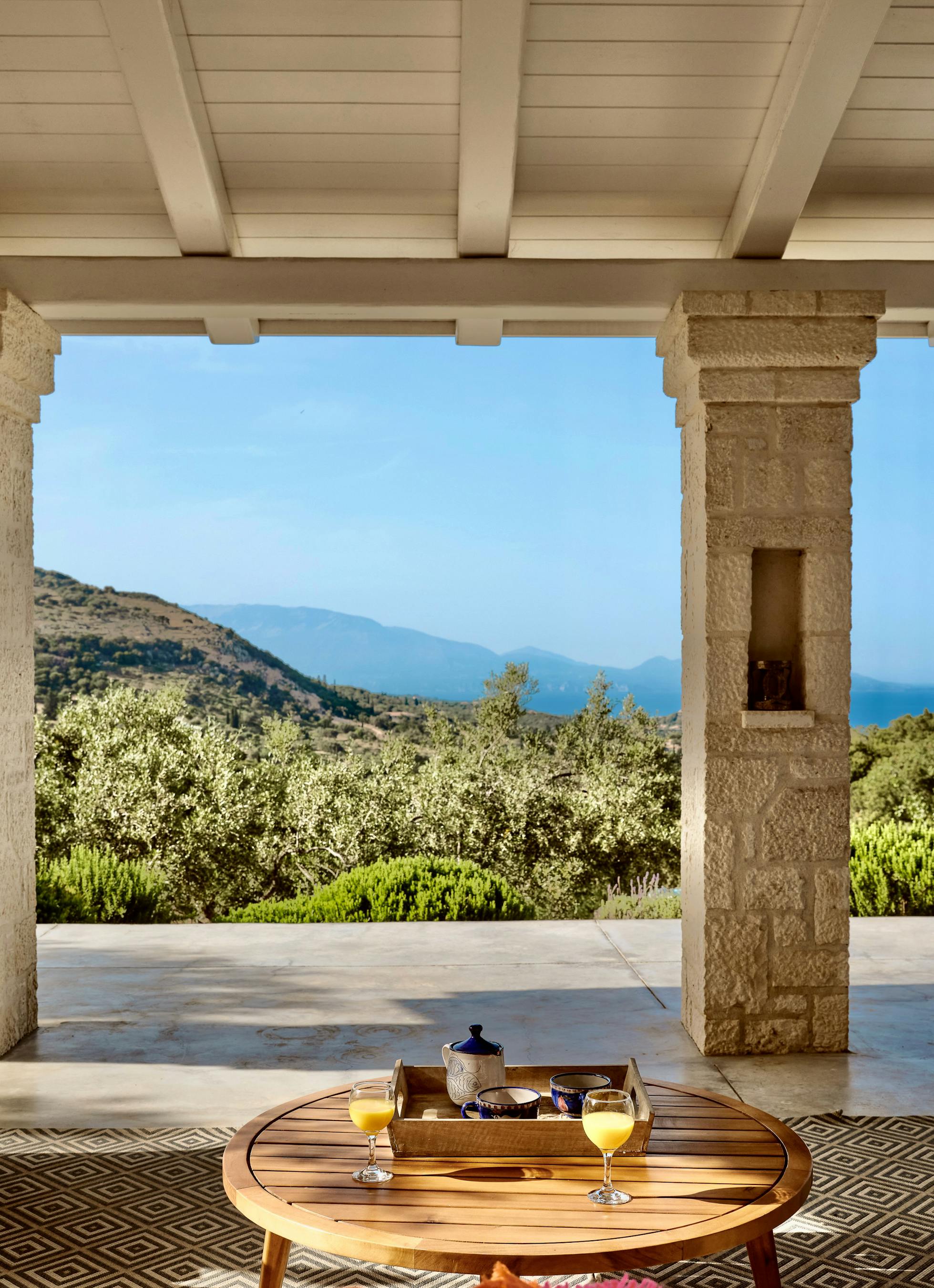 Covered terrace with stone columns and exposed beam ceiling framing panoramic views of olive groves and distant coastline. Teak lounge furniture with turquoise cushions arranged around round coffee table set for breakfast.