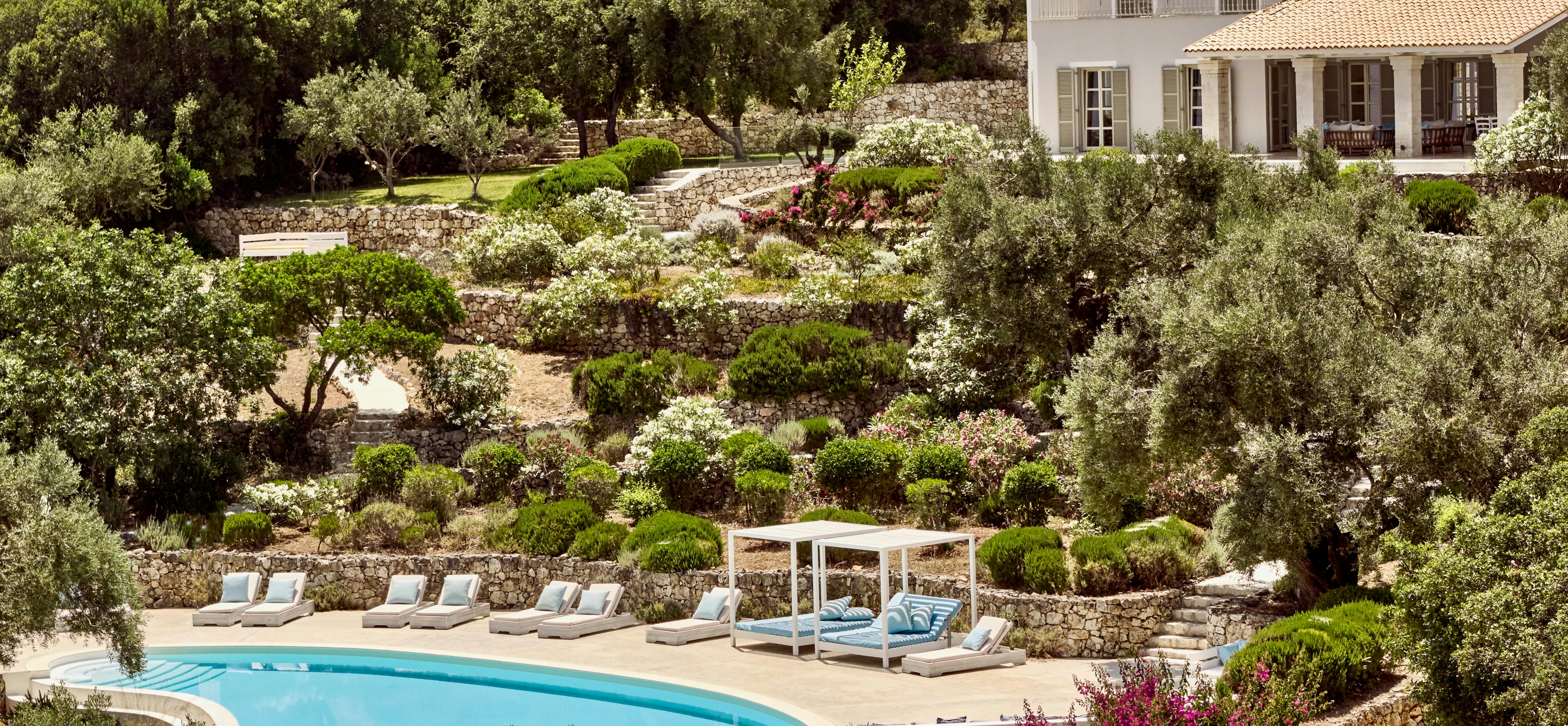 Hillside estate with white villa overlooking terraced gardens and curved pool below. Stone retaining walls and mature olive trees frame the Mediterranean landscape setting.