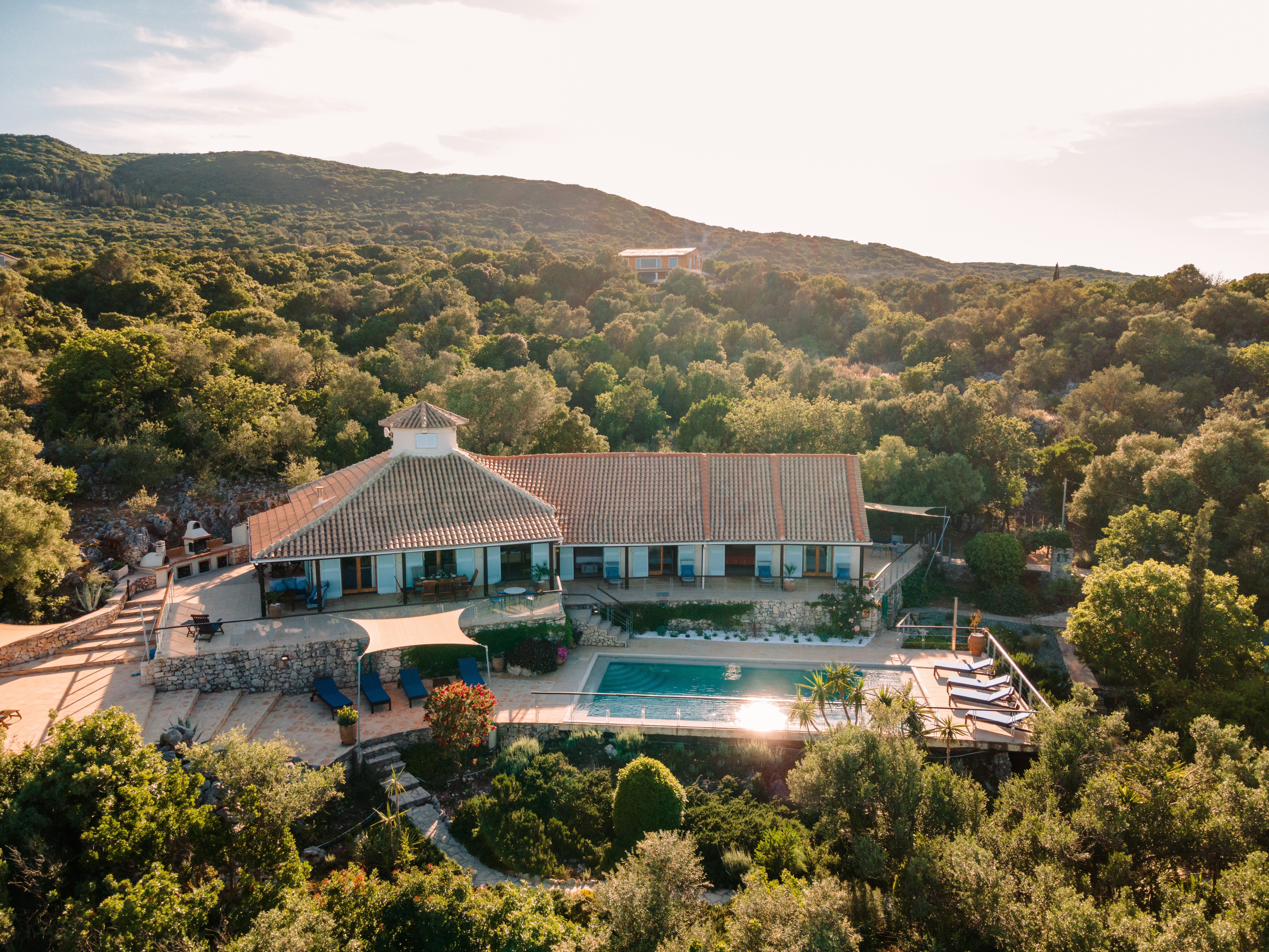 An aerial view captures a white Mediterranean villa with a central turret nestled in dense olive groves on a hillside, with terraced pool areas and mountain views in the distance.