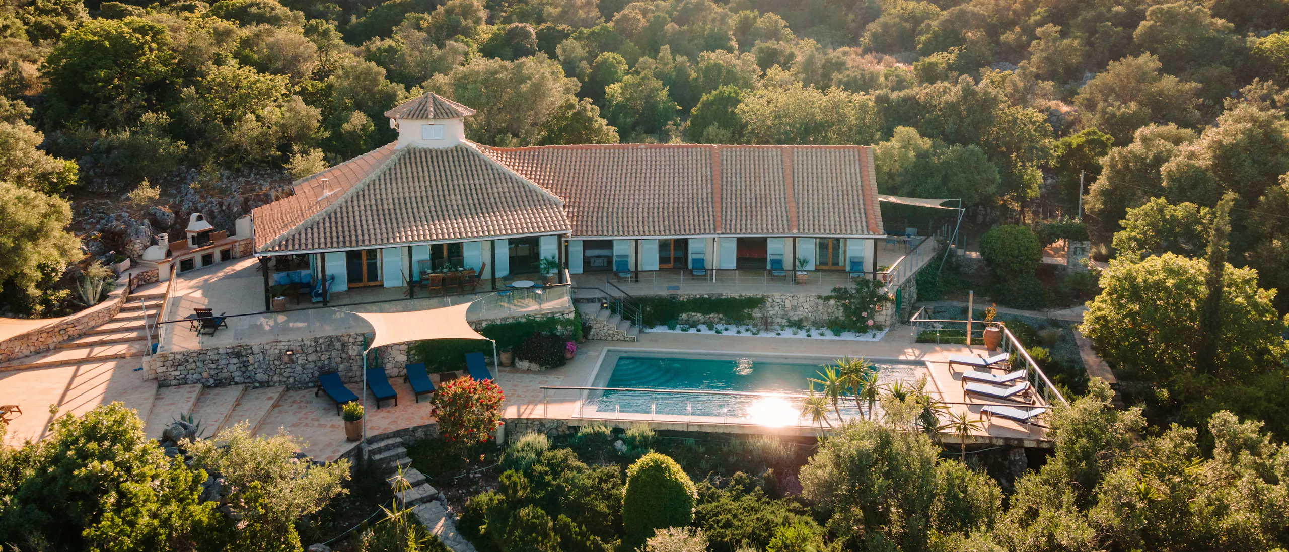 An aerial view captures a white Mediterranean villa with a central turret nestled in dense olive groves on a hillside, with terraced pool areas and mountain views in the distance.