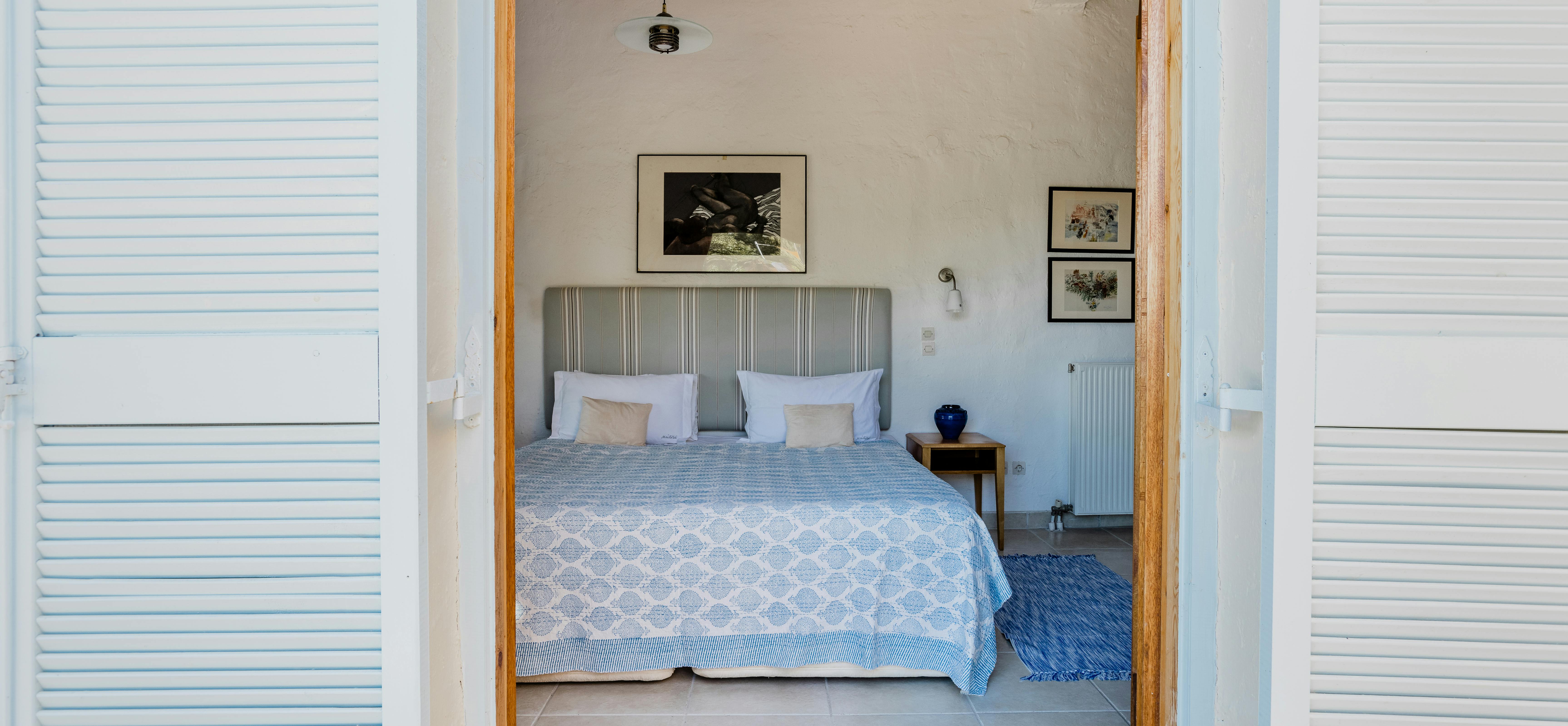 A bedroom viewed through open white louvered shutters displays a striped grey headboard, blue patterned bedding, and simple coastal decor.