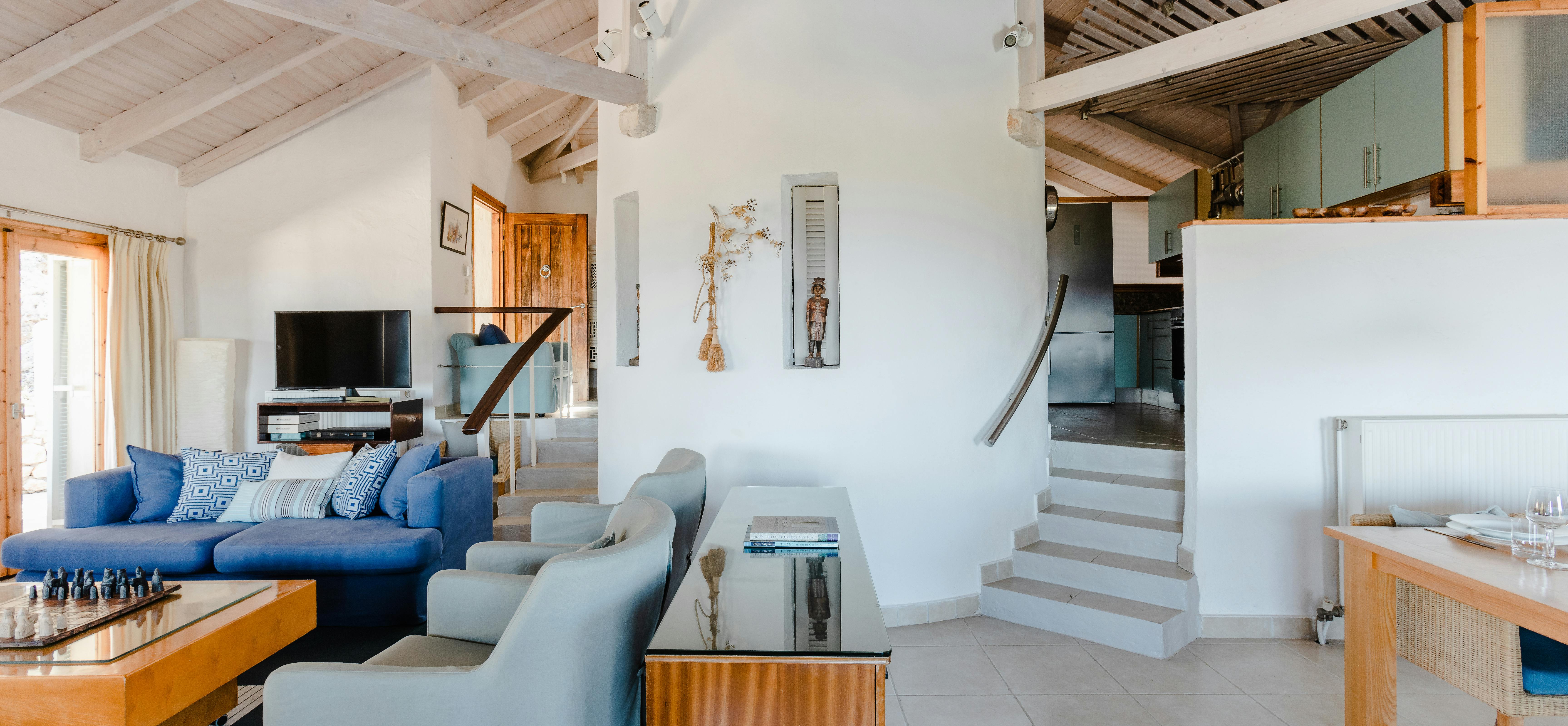 An open-plan living space showcases a white cylindrical column supporting a mezzanine level, with blue upholstered furniture, wooden accents, and stairs leading to the kitchen area above.