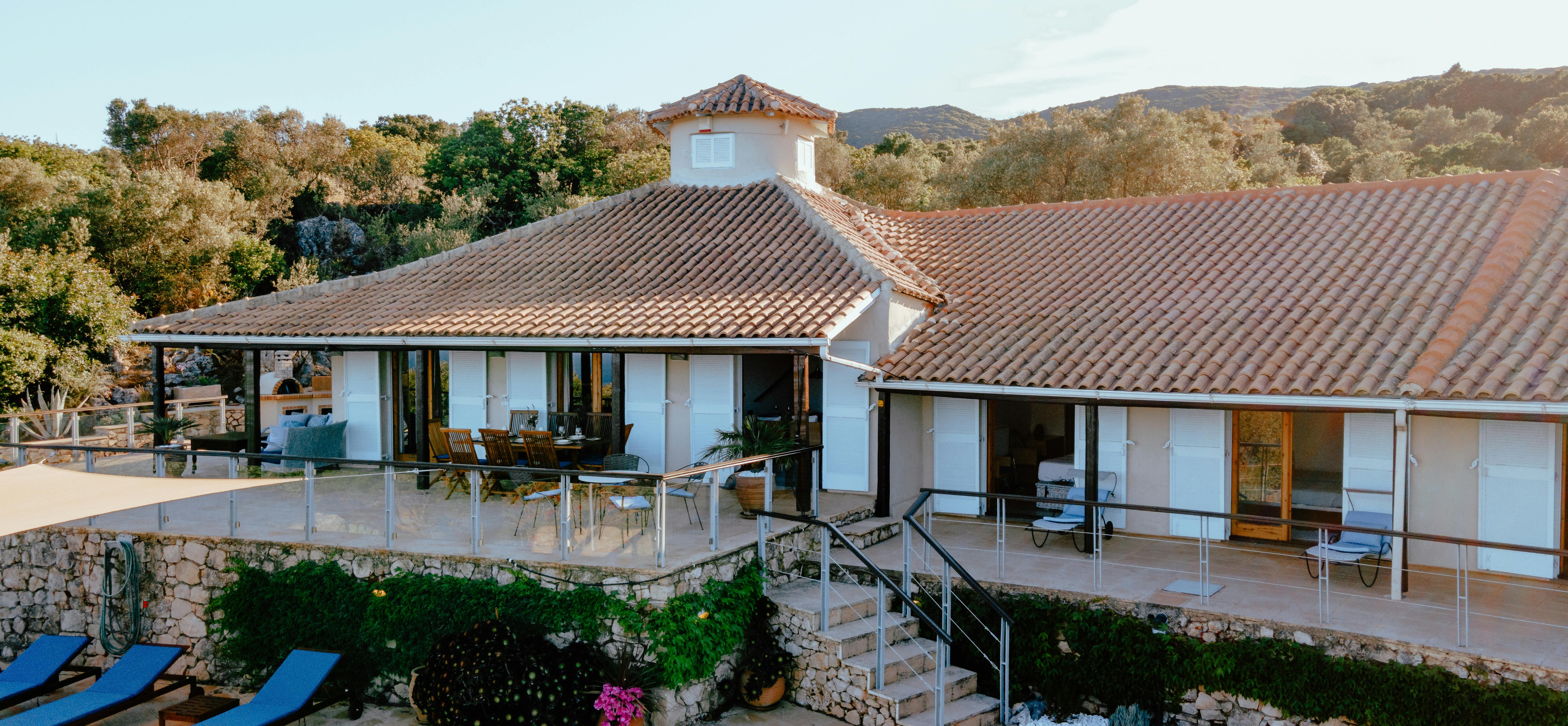The villa's two-level facade at dusk shows terracotta tile roofing, multiple covered terraces with glass railings, and the illuminated pool area surrounded by landscaped gardens.