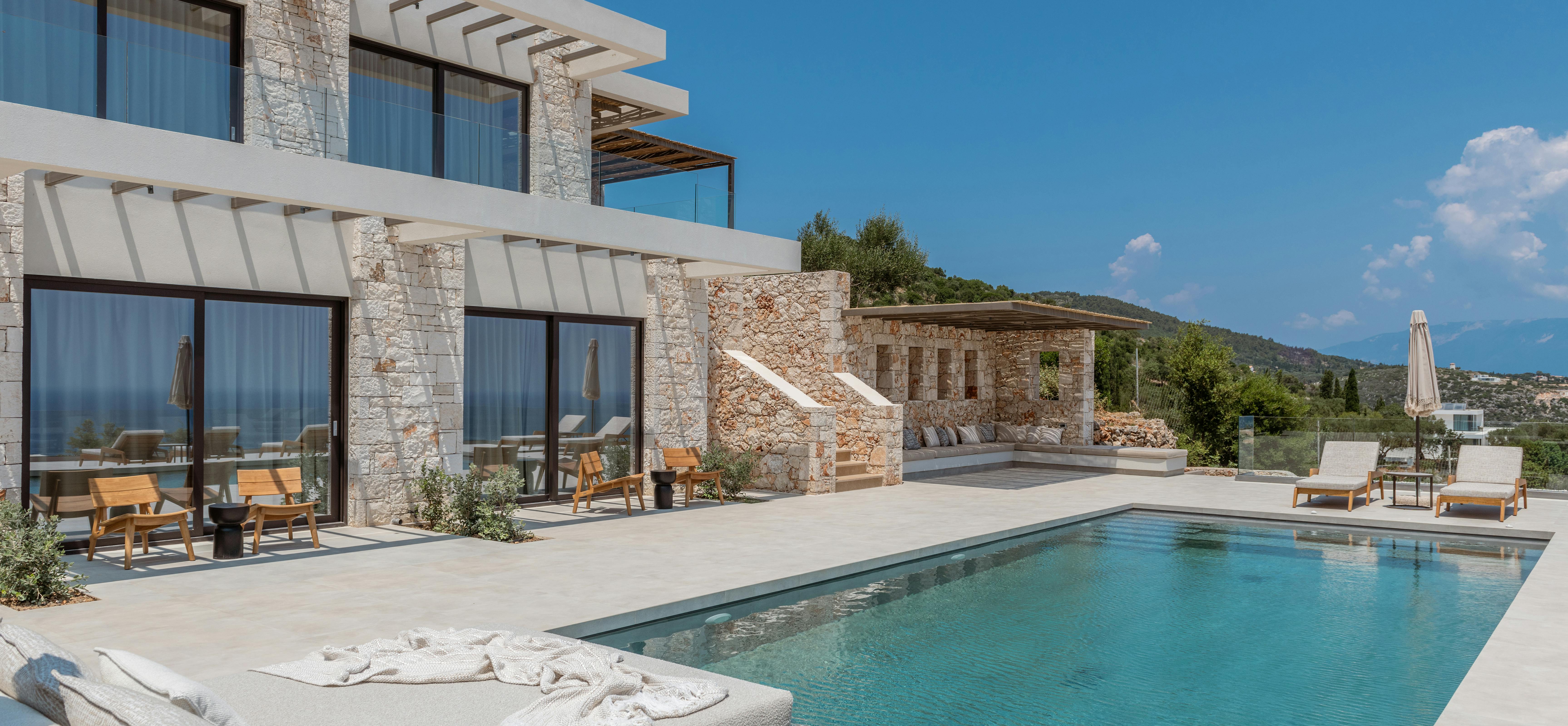 The multi-level villa exterior showcases an infinity pool, textured stone walls, a wooden pergola structure, and panoramic views of the sea and distant islands.
