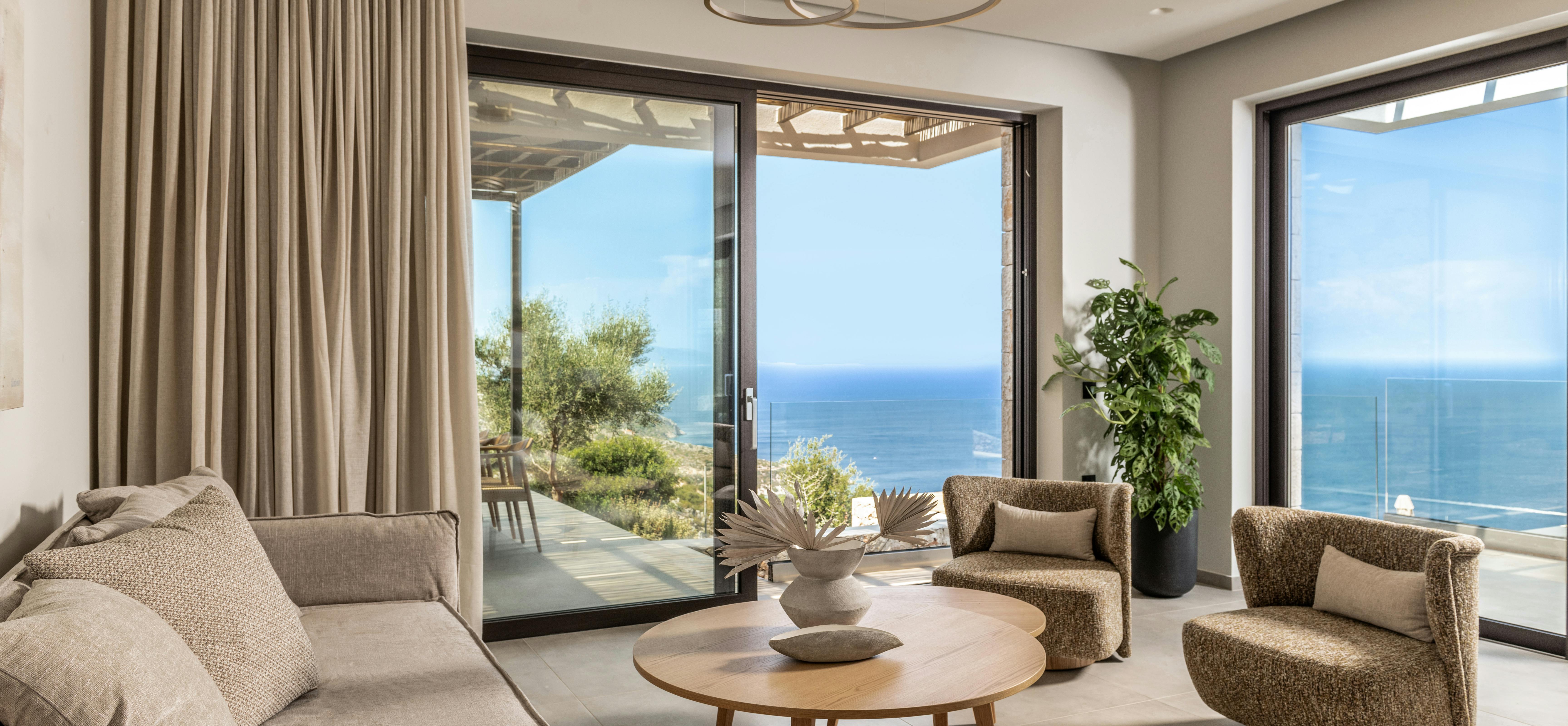 A modern coastal living room features neutral-toned furniture, circular brass pendant lights, and floor-to-ceiling windows offering sweeping ocean views.