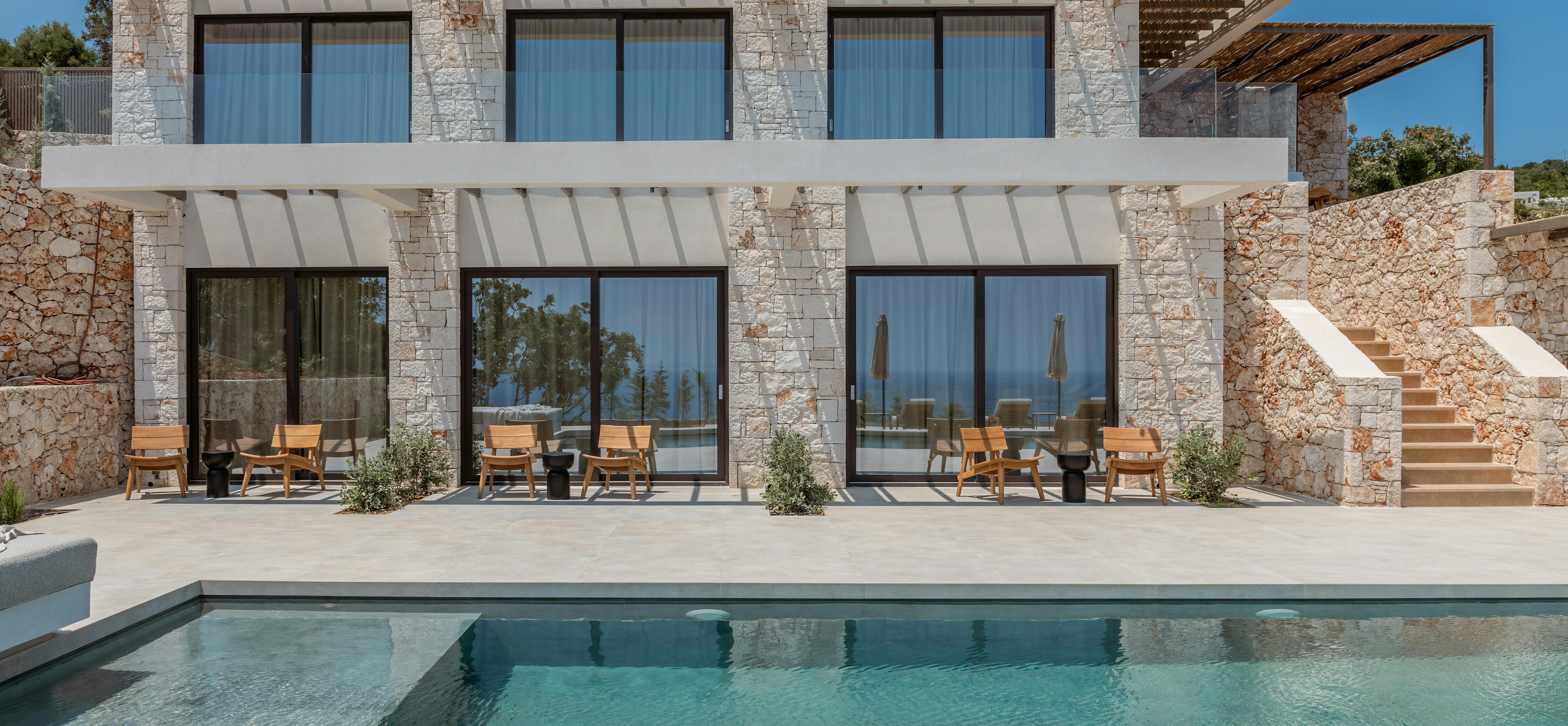 The two-story villa facade displays stone columns, white pergola overhangs on both levels, and a ground-level infinity pool stretching across the terrace.