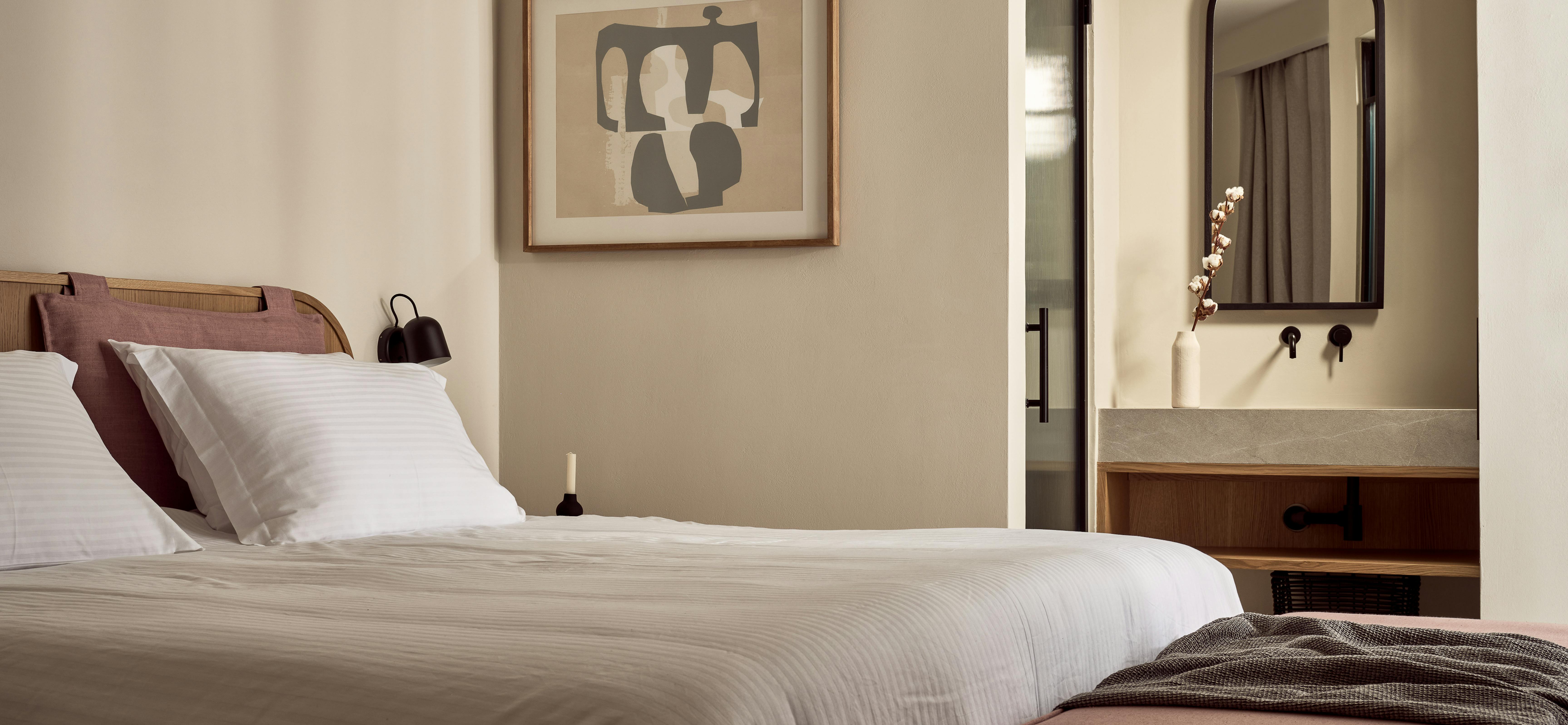 A serene bedroom with cane-backed headboard, abstract artwork, white bedding with mauve accents, and arched doorway to an ensuite bathroom.