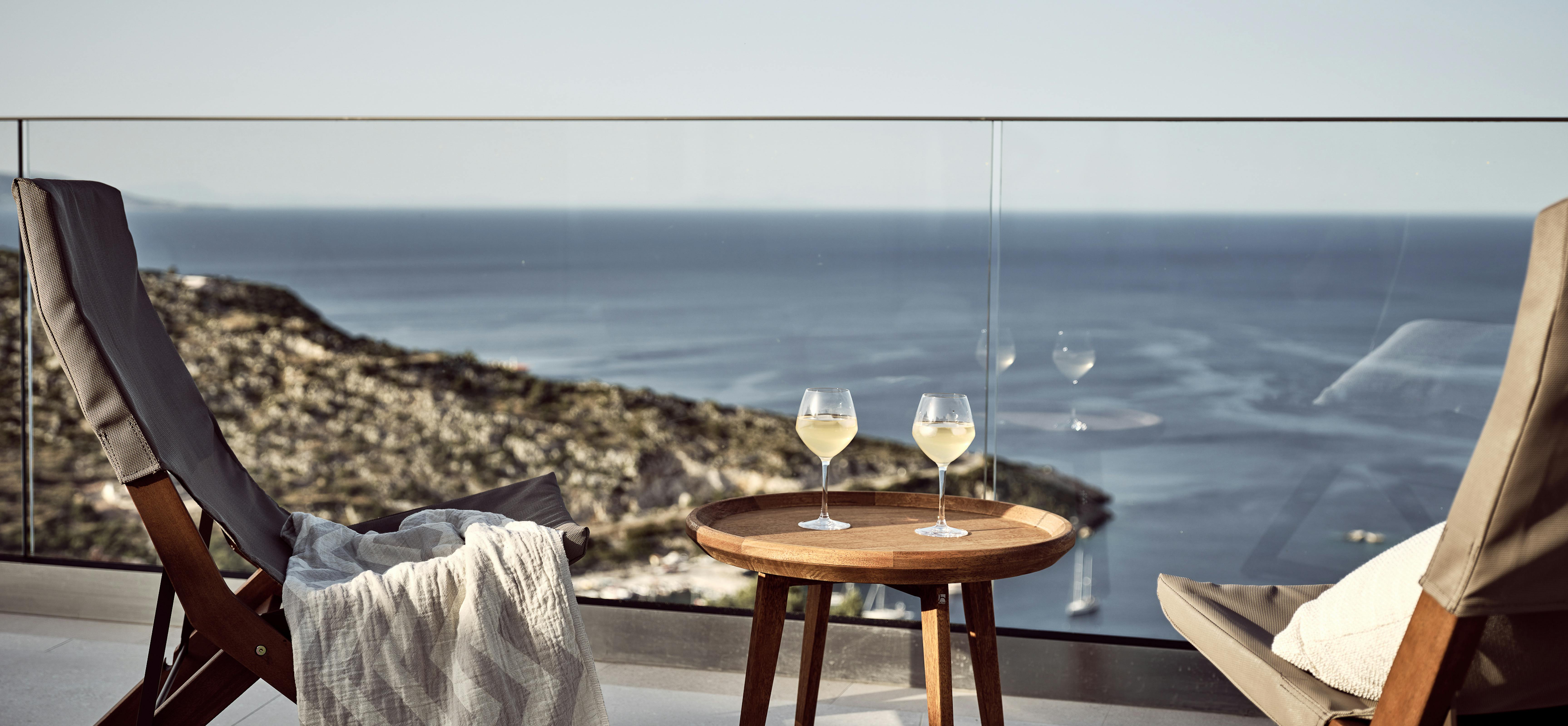 Two wooden deck chairs on a terrace with wine glasses on a side table, glass railing, and panoramic coastal views.