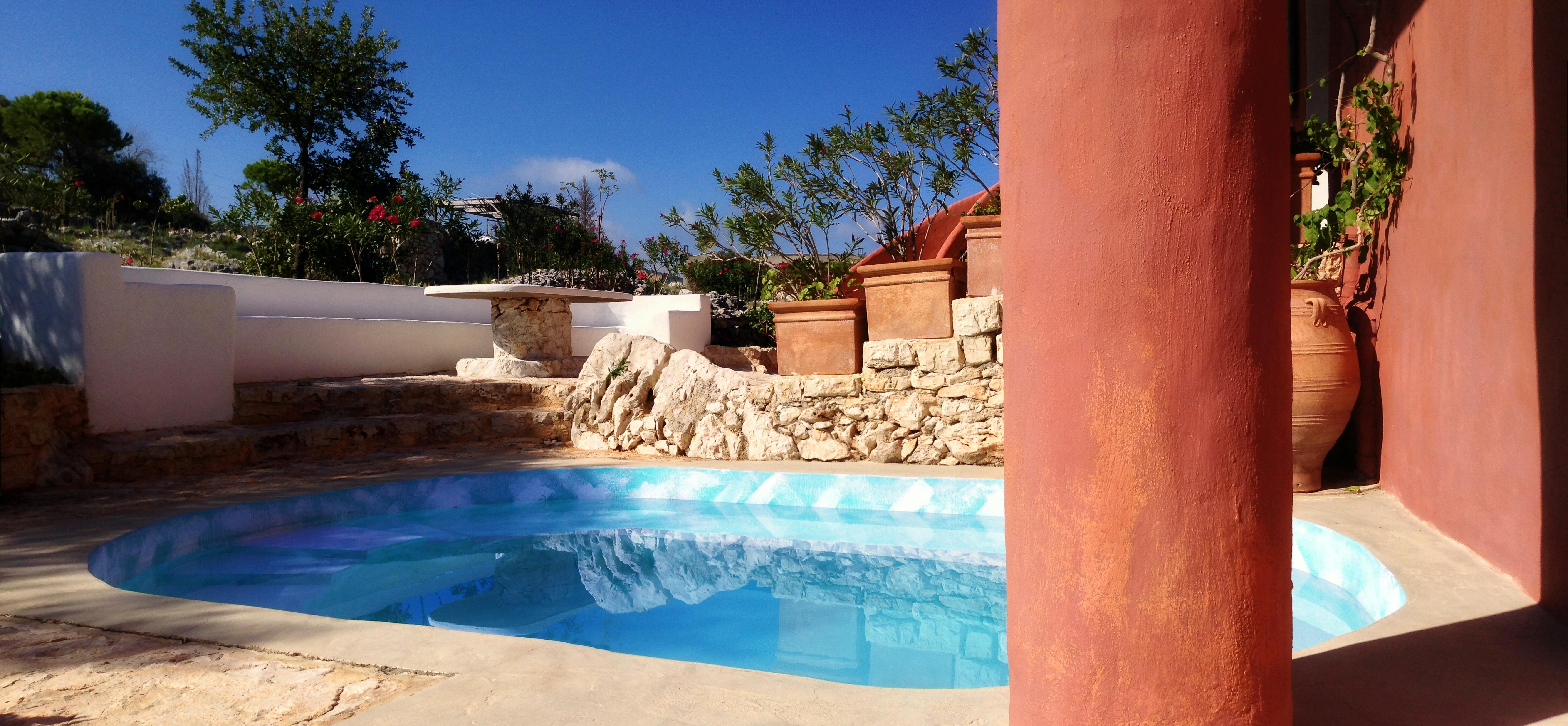 A curved turquoise swimming pool with stone paving surrounded by coral-pink stucco walls, terracotta planters with small trees and flowering plants, stone retaining walls, and bright blue Mediterranean sky.
