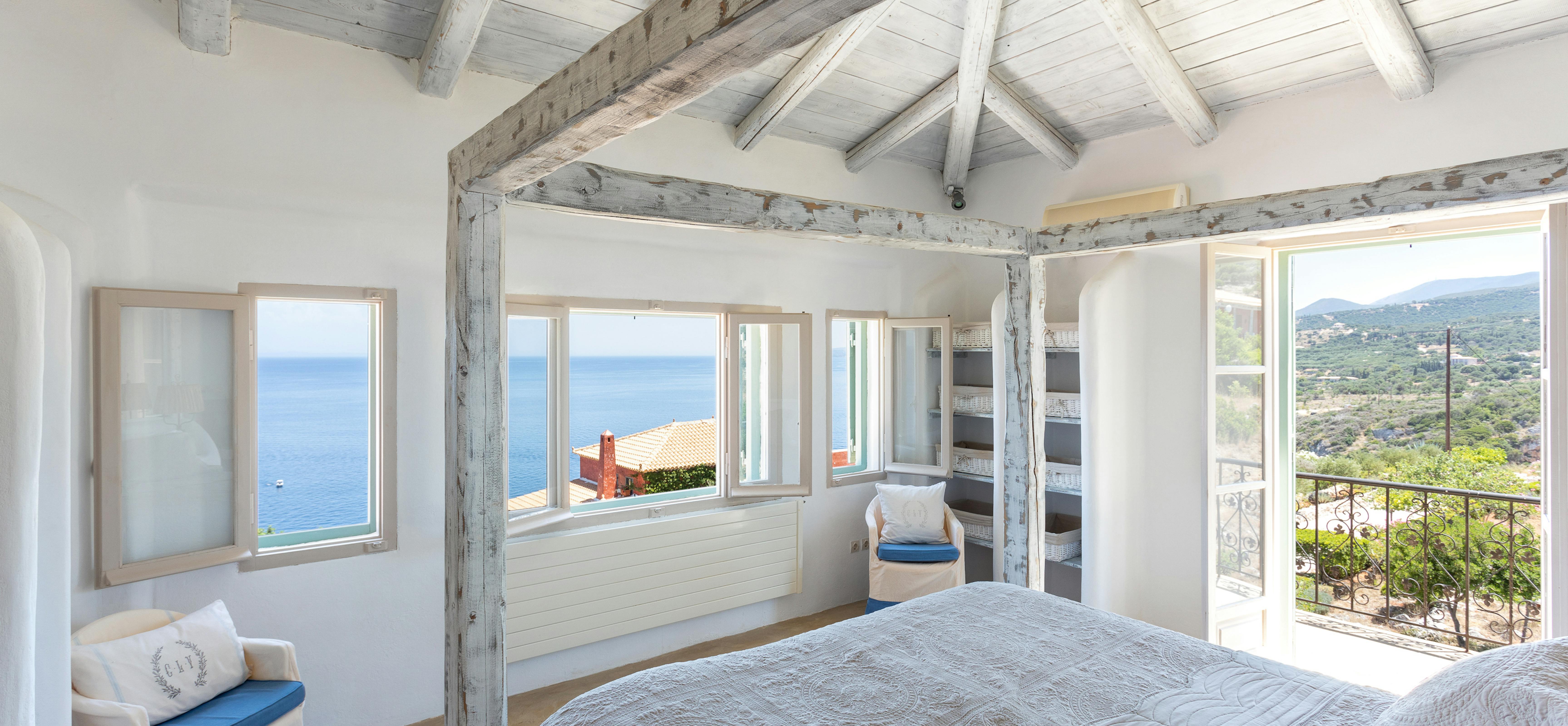 A coastal bedroom with whitewashed wood plank ceiling and exposed beams, grey quilted bedding, multiple windows framing ocean and hillside views, built-in shelving, white chairs with blue cushions, and natural light.