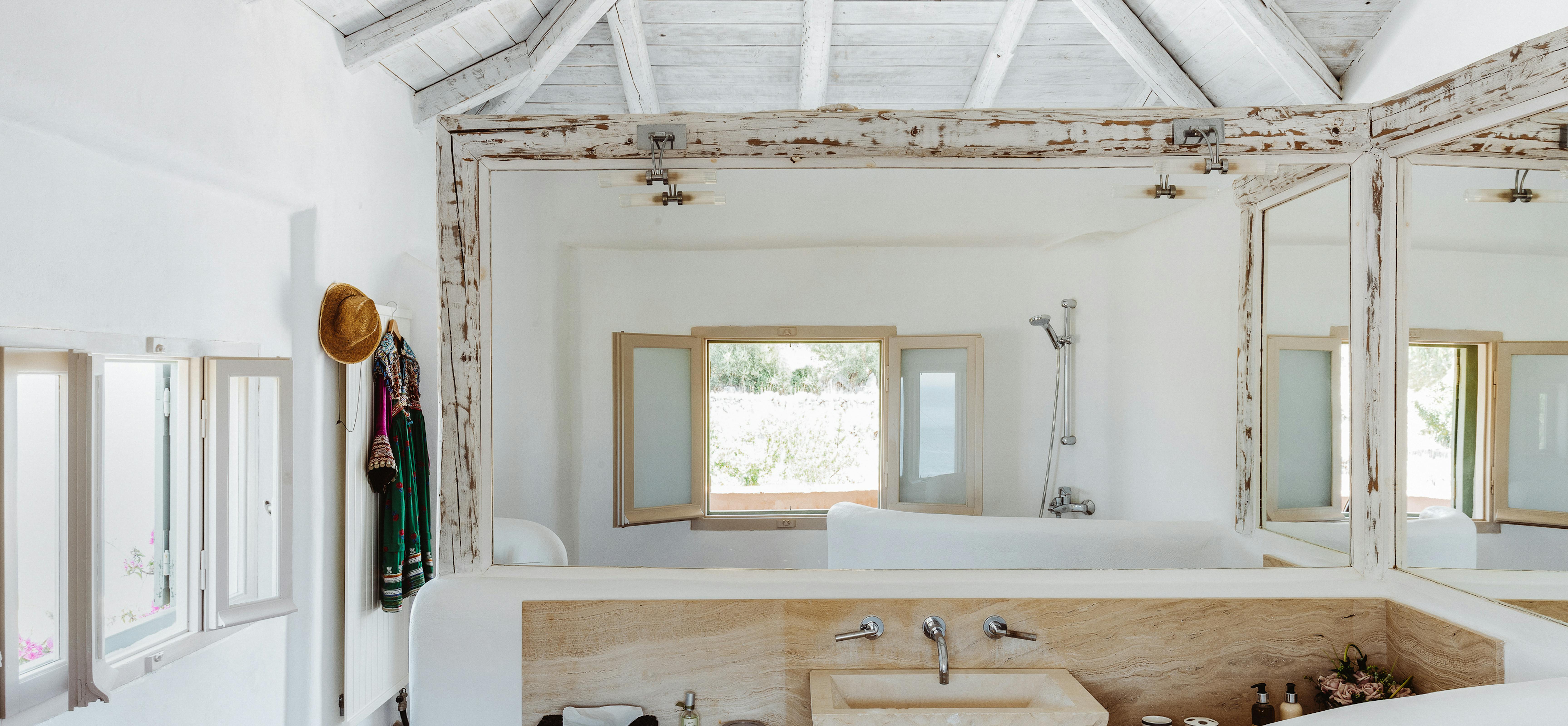A luxurious open bathroom with whitewashed exposed beam ceiling, freestanding white tub, carved stone vessel sink on wood counter, tripod table lamps, colourful garment hanging on hooks, and windows with ocean and garden views.