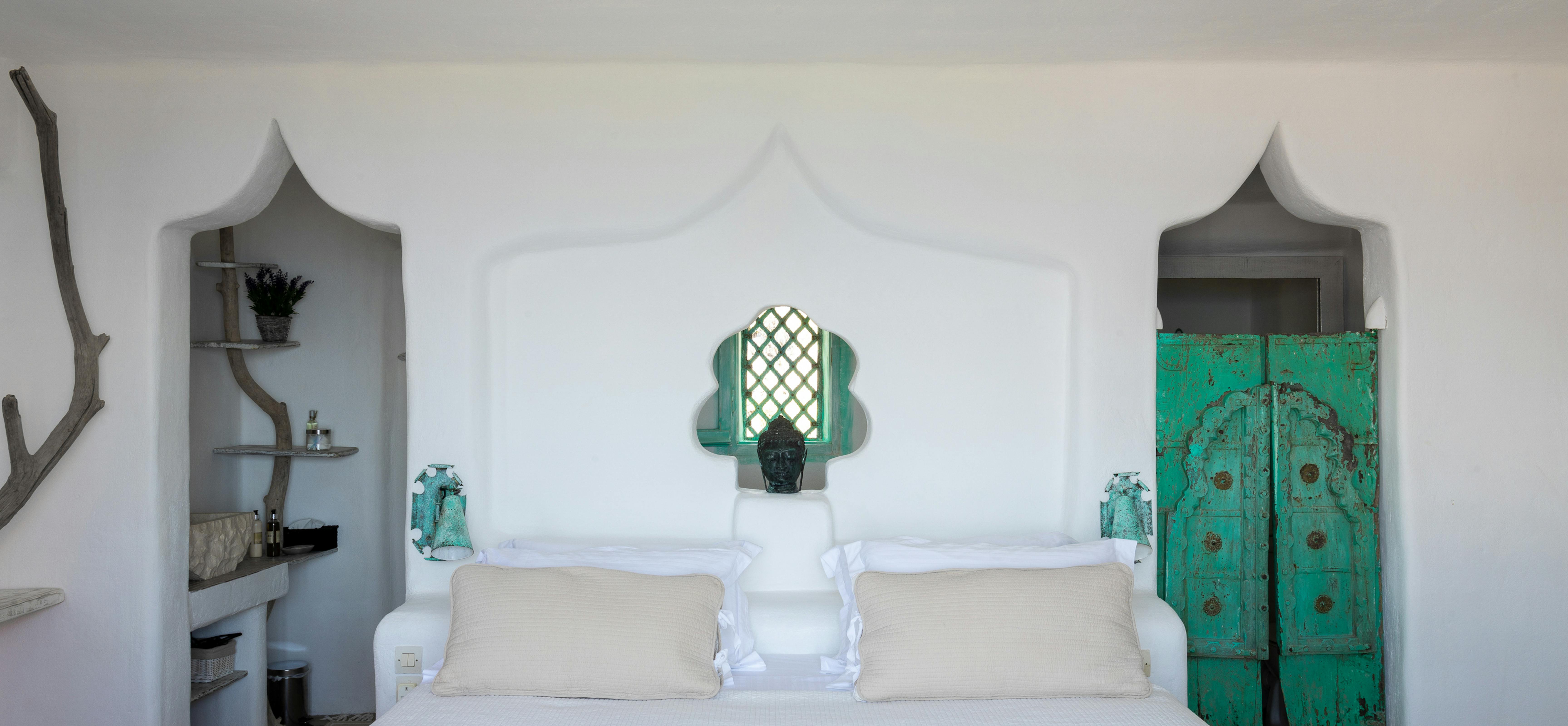 A minimalist all-white bedroom with symmetrical design featuring a bed with cream linens, two Moroccan-style arched alcove niches flanking the headboard, distressed turquoise vintage doors as nightstands, and decorative skull sculpture.