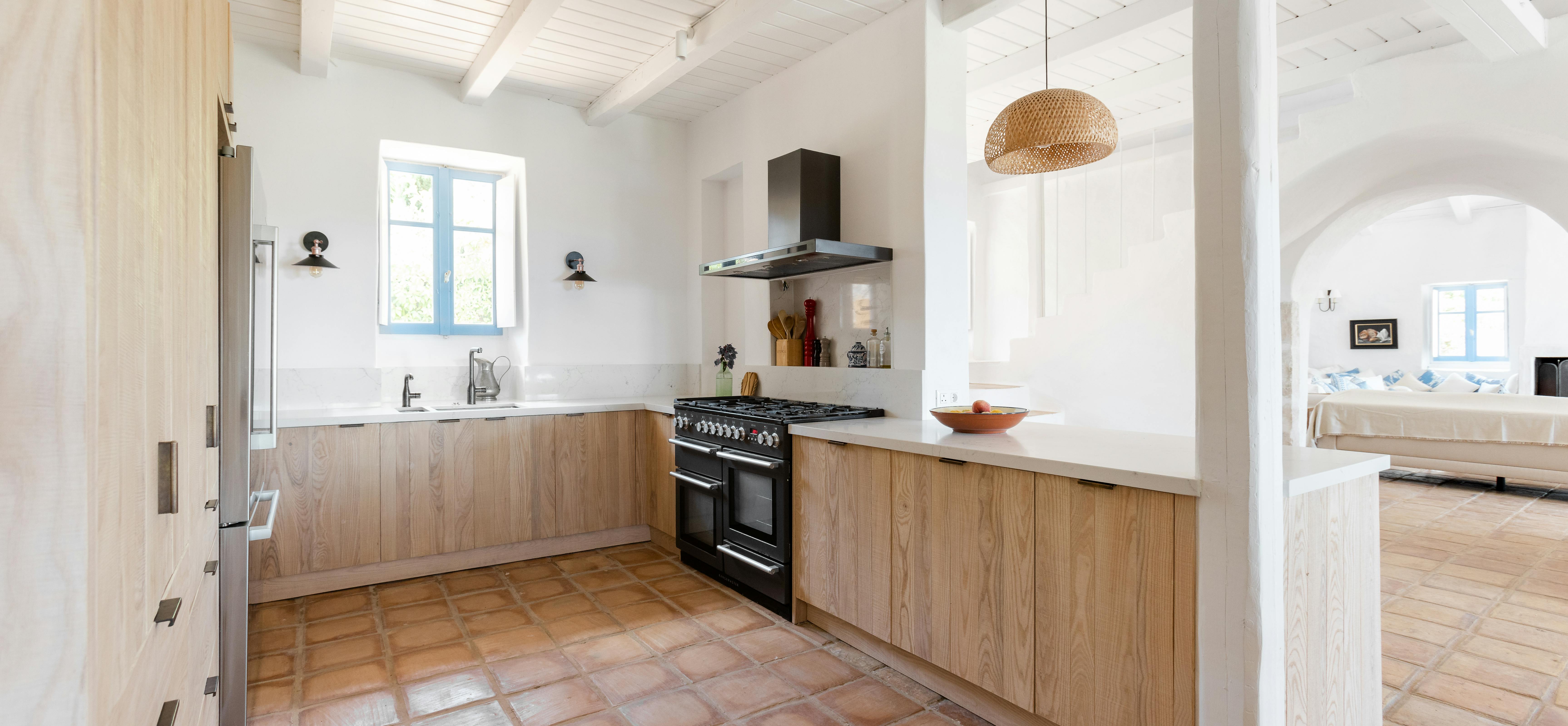 A modern Mediterranean kitchen with light wood cabinetry, black range cooker, terracotta tile floor, woven pendant light, and white walls with exposed beams.